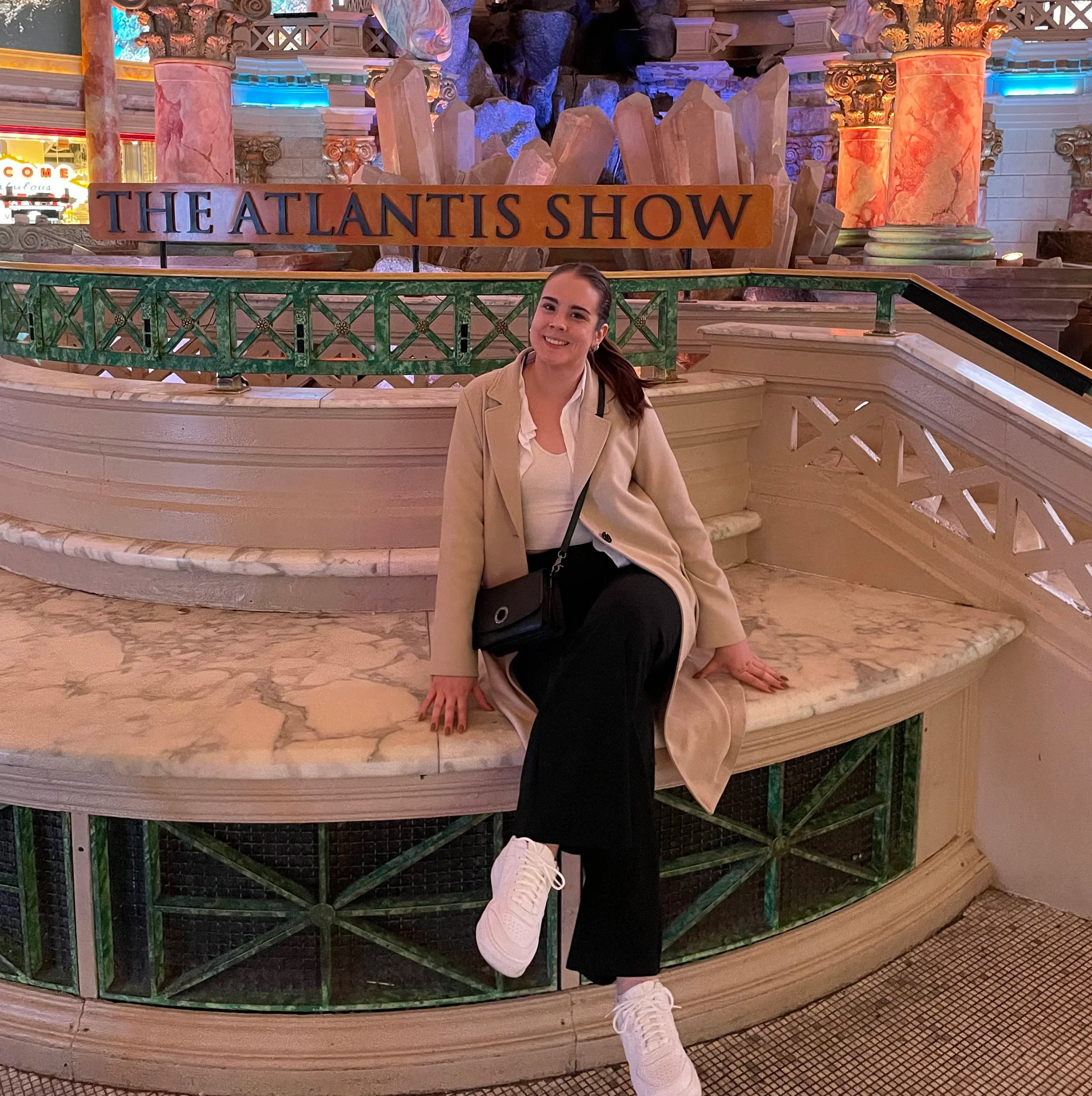 A young woman with dark hair and a beige blazer sits on the floor, smiling at the camera, in front of the display sign for The Atlantis Show at an indoor venue. Part of another person's shoe is visible at the bottom of the image. The background features decorative columns and a rocky, blue-lit centerpiece.