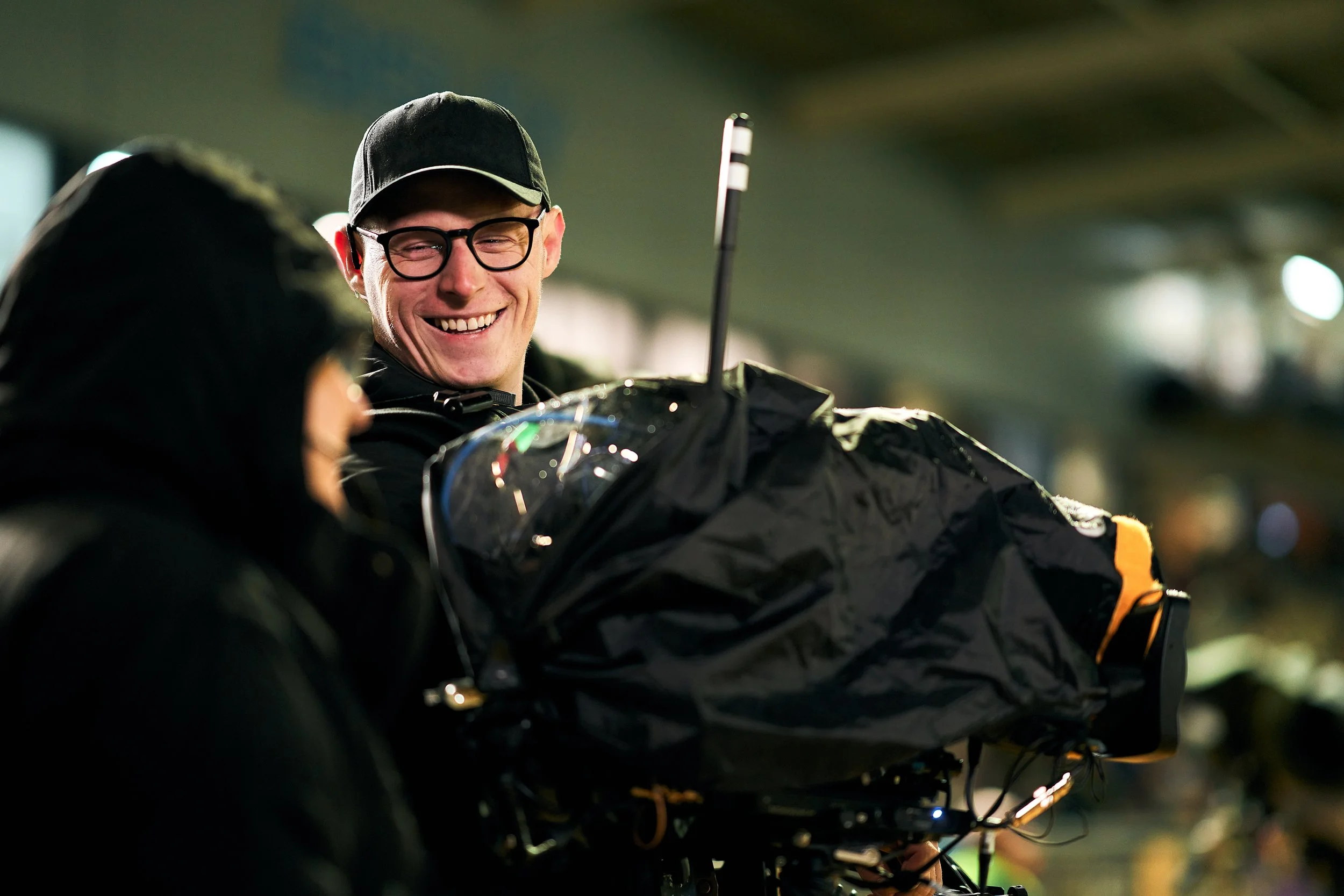 A man smiling with glasses and a black cap talking to another person in a dark jacket inside a well-lit building.