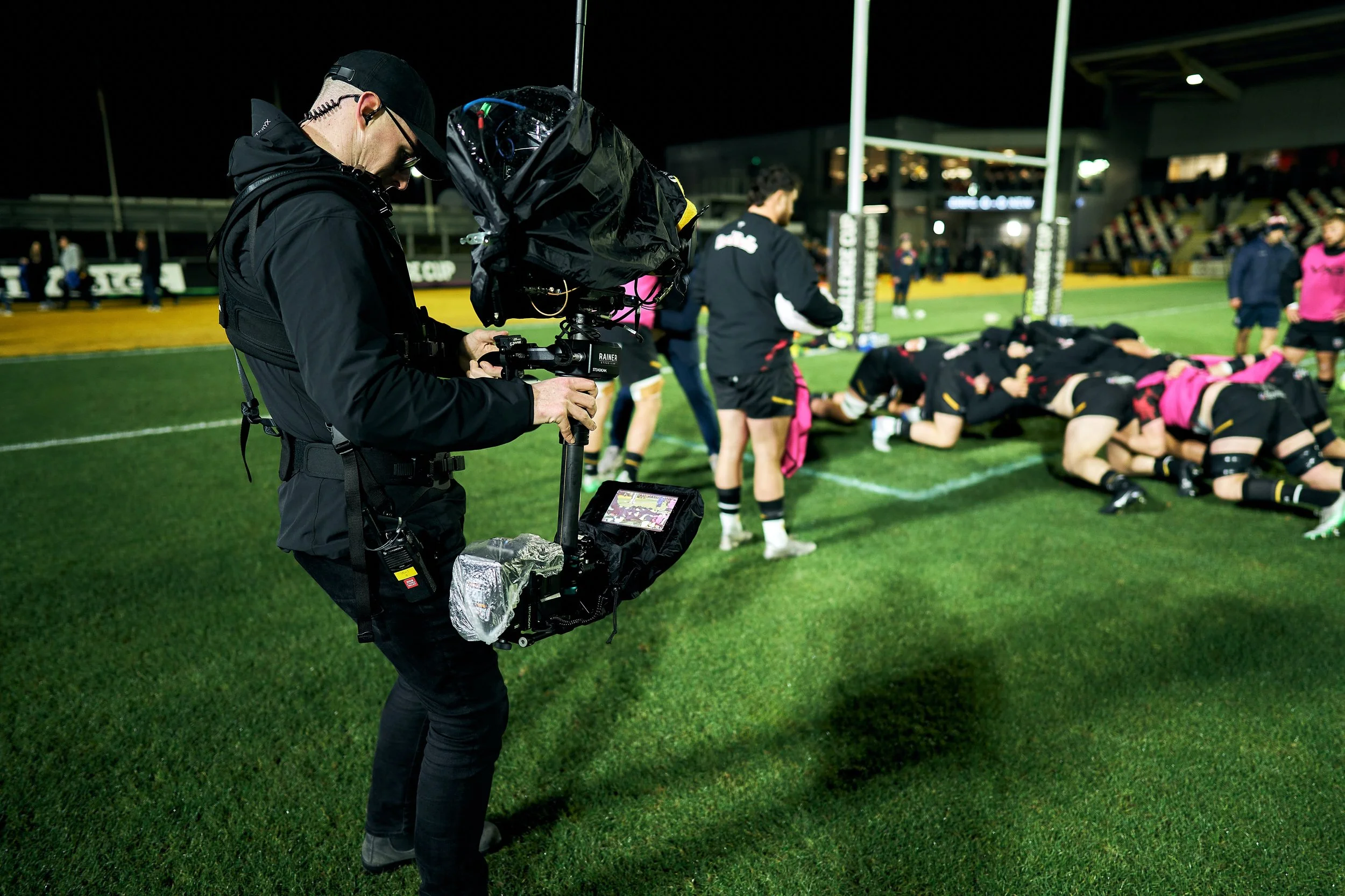 A videographer filming a rugby game on a field at night, with players in a scrum in the background.