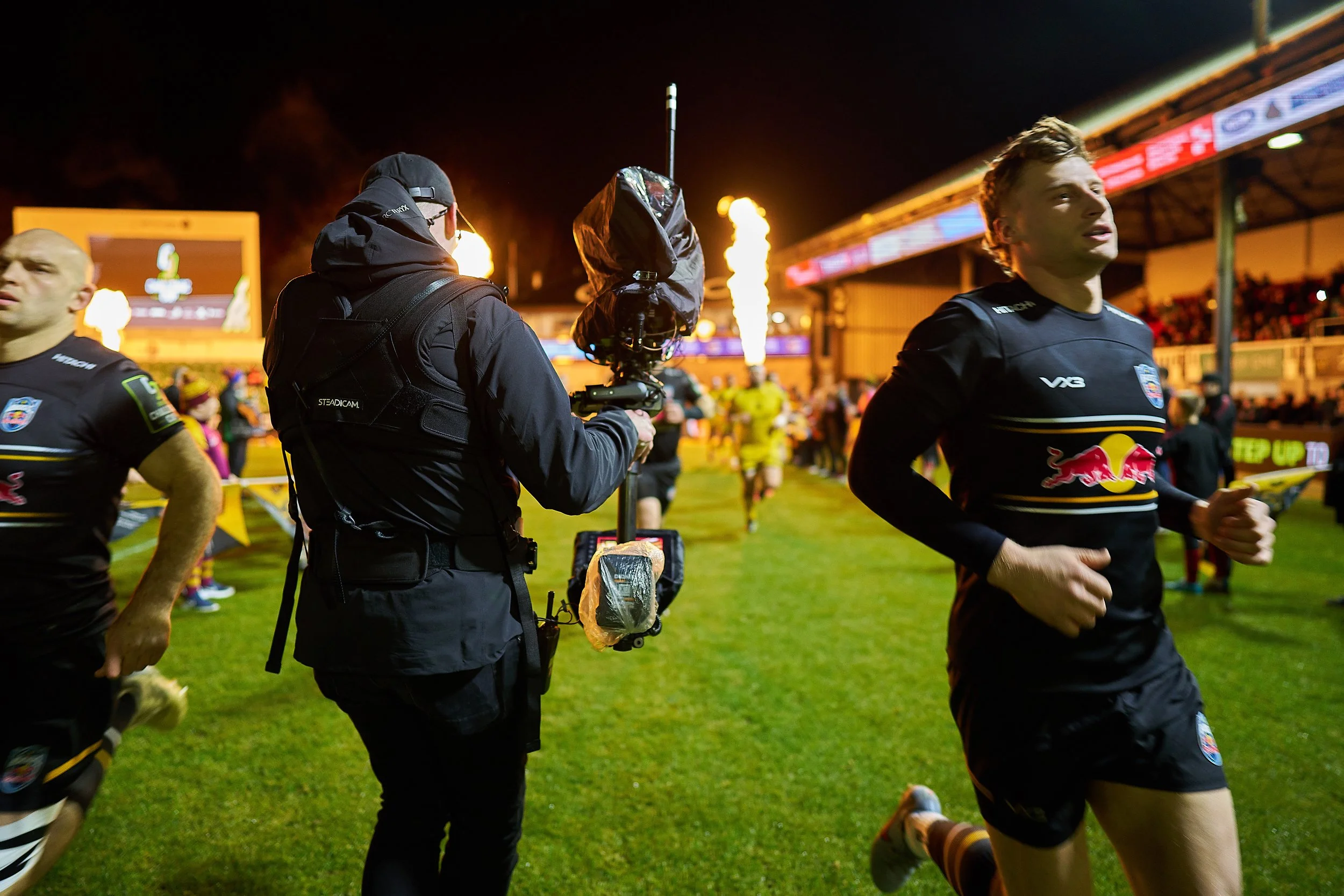 Rugby players warming up on a field during a nighttime game with bright flames and stadium lights in the background, and a cameraman capturing footage.