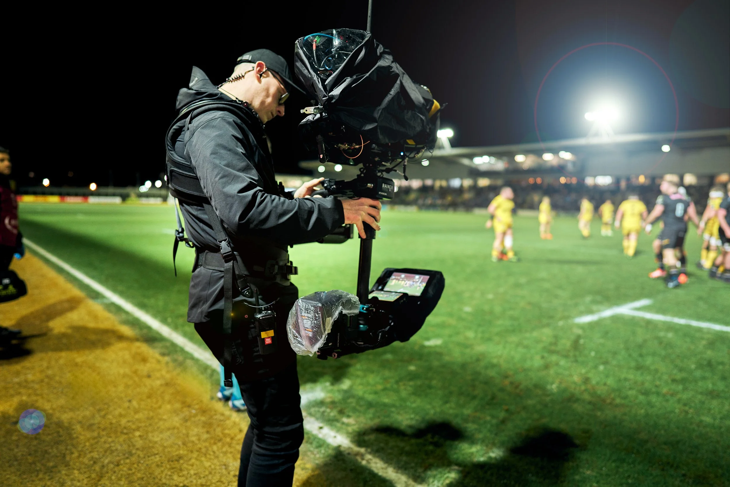 A camera operator filming a nighttime soccer match on a brightly lit field, with players in yellow and black uniforms visible in the background.
