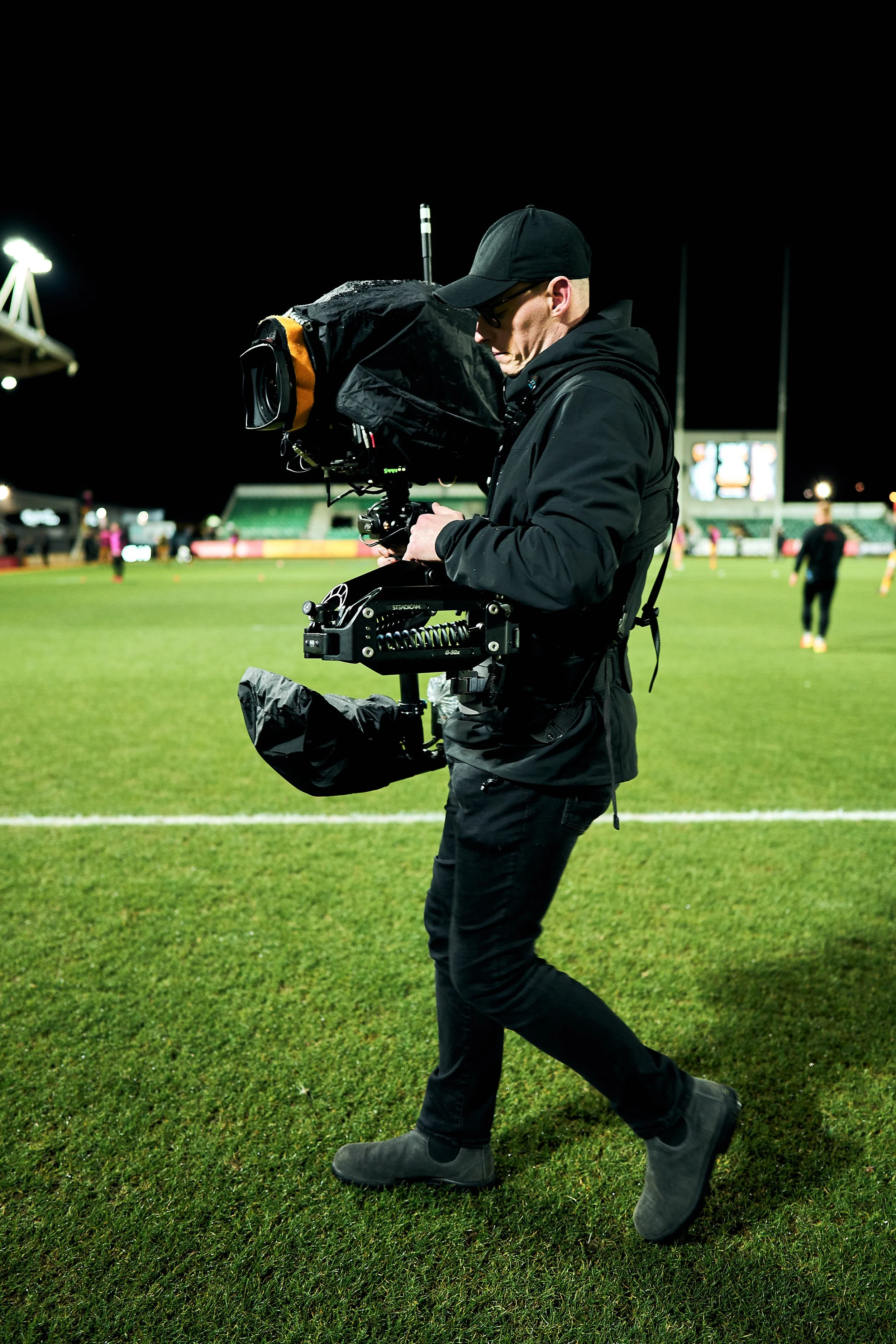 A videographer in black clothing and cap filming on a grassy field at night with stadium lights and football players in the background.