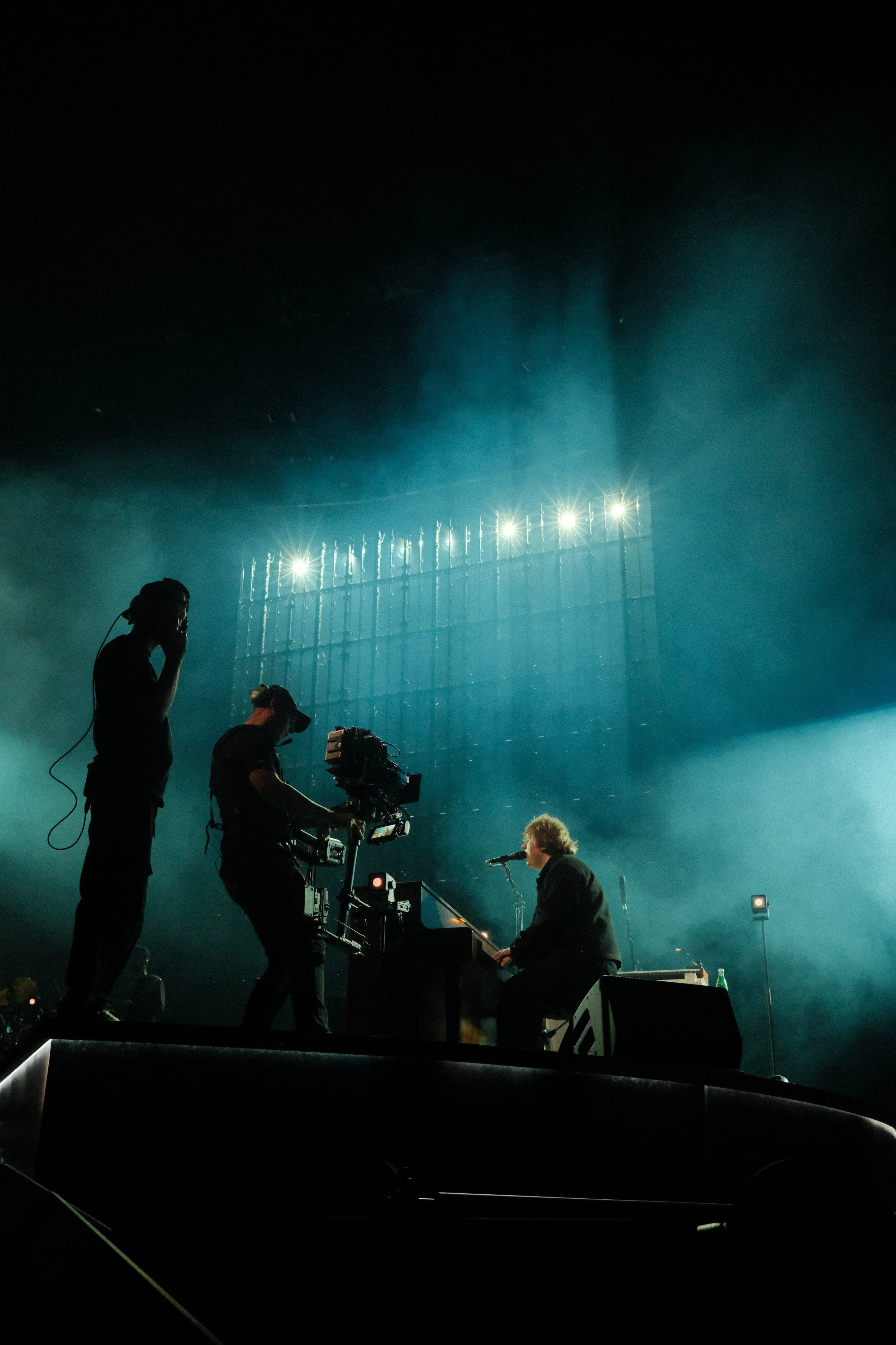 A musician playing keyboard on stage with camera crew filming him at night under bright stage lights with smoke effects.