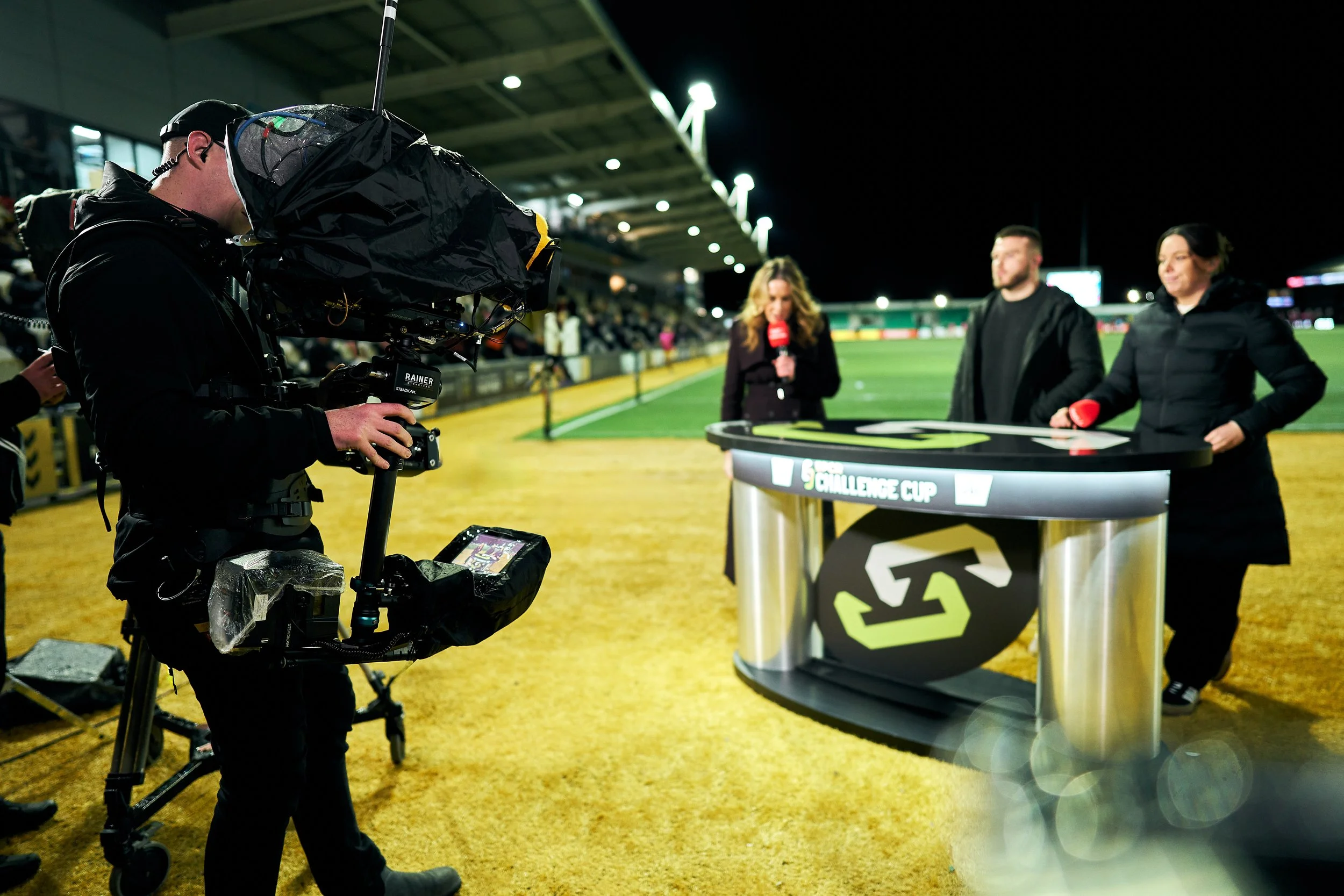 Nighttime sports broadcast with two female hosts and a male guest at a stadium. A cameraman films the scene with a professional camera. The broadcast table has a logo and is labeled 'Challenge Cup'. The stadium seating and field are visible in the ba