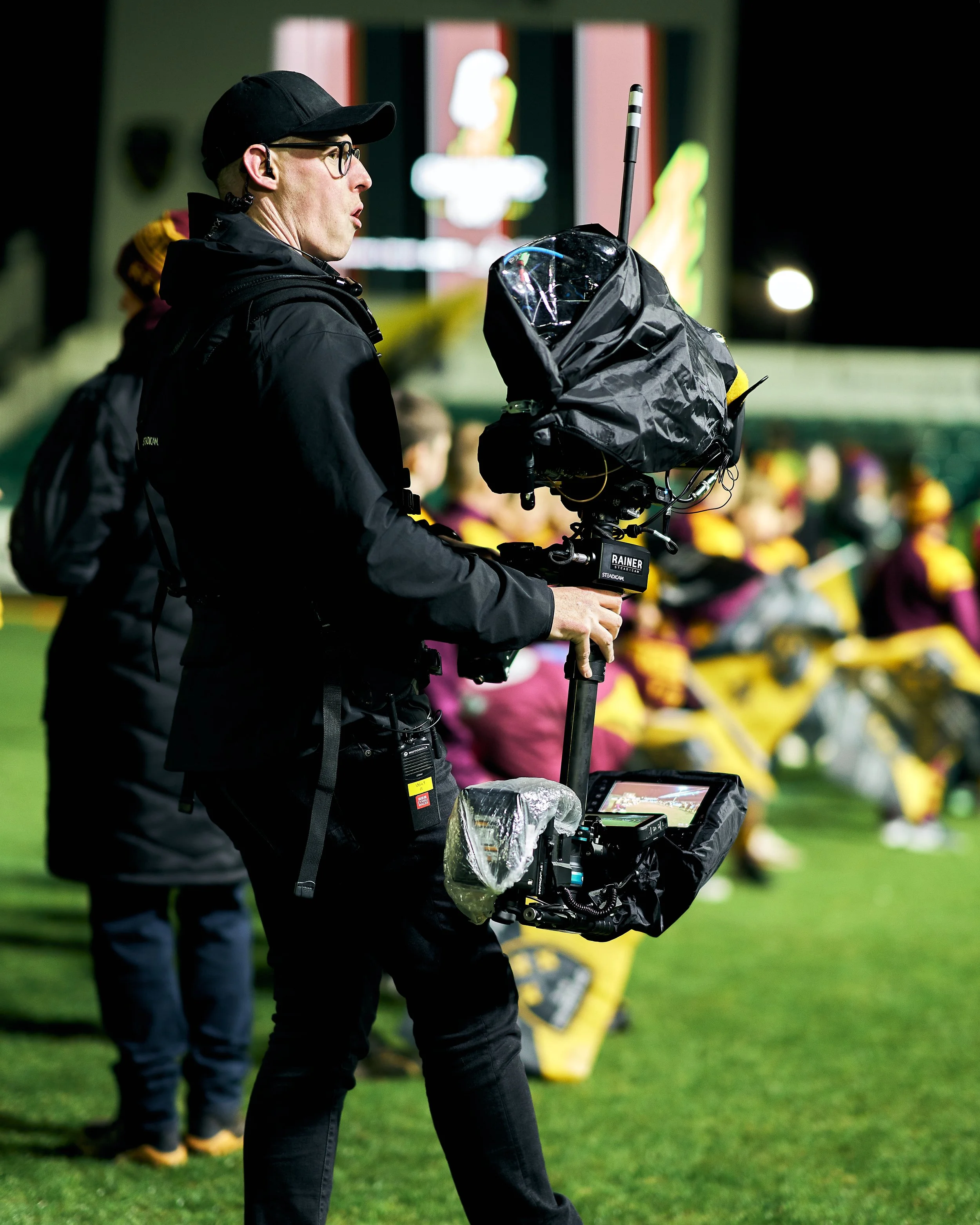 A cameraman filming a sports event at night, with a professional camera stabilizer, on a field with people sitting in the background.