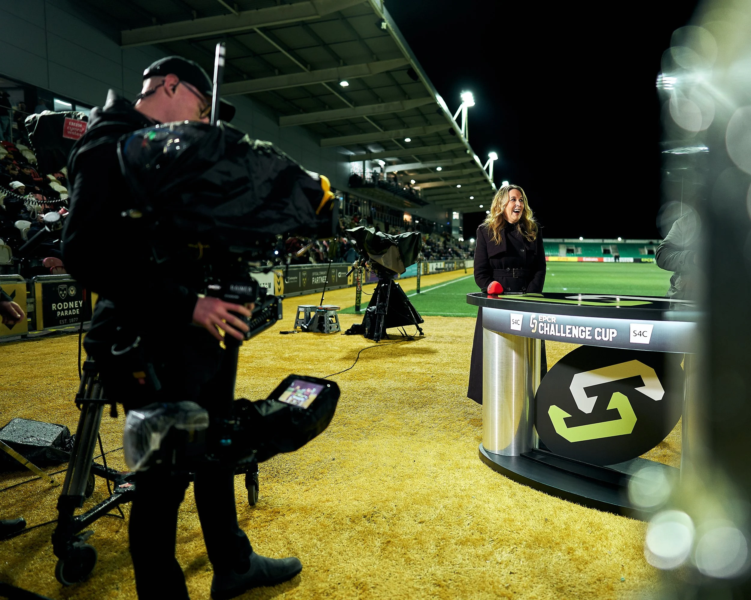 A woman is being filmed by a cameraman during a nighttime broadcast at a sports stadium. She is standing at a table with a sign that reads 'EPCR Challenge Cup' and is smiling.