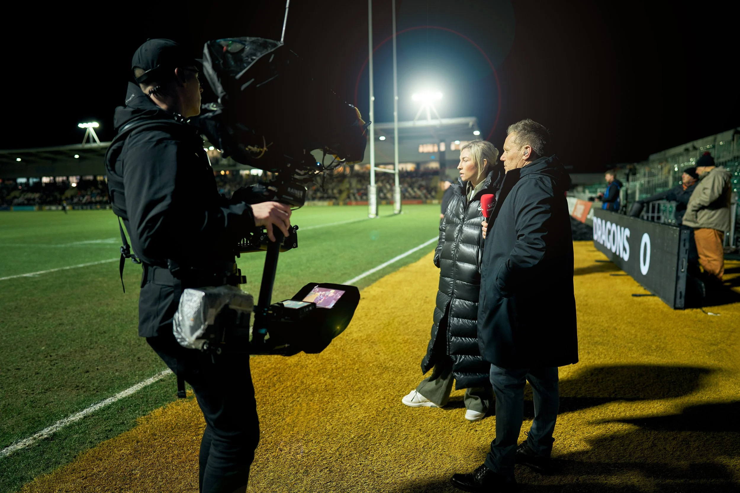 Two sports interviewers, a woman and a man, speak into microphones while being filmed at a rugby stadium at night.