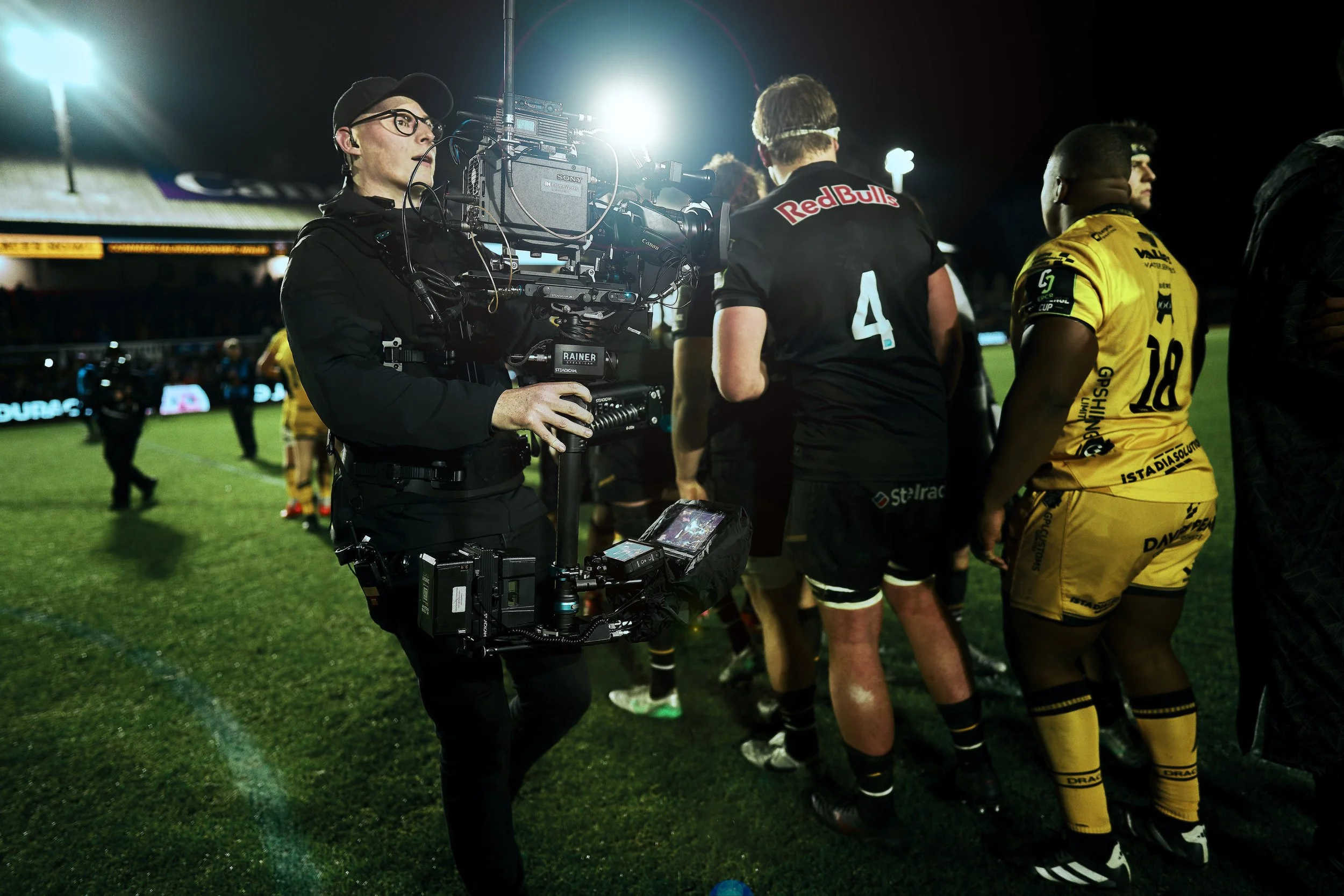 Camera operator filming a rugby match at night, with players in black and yellow uniforms on the field.
