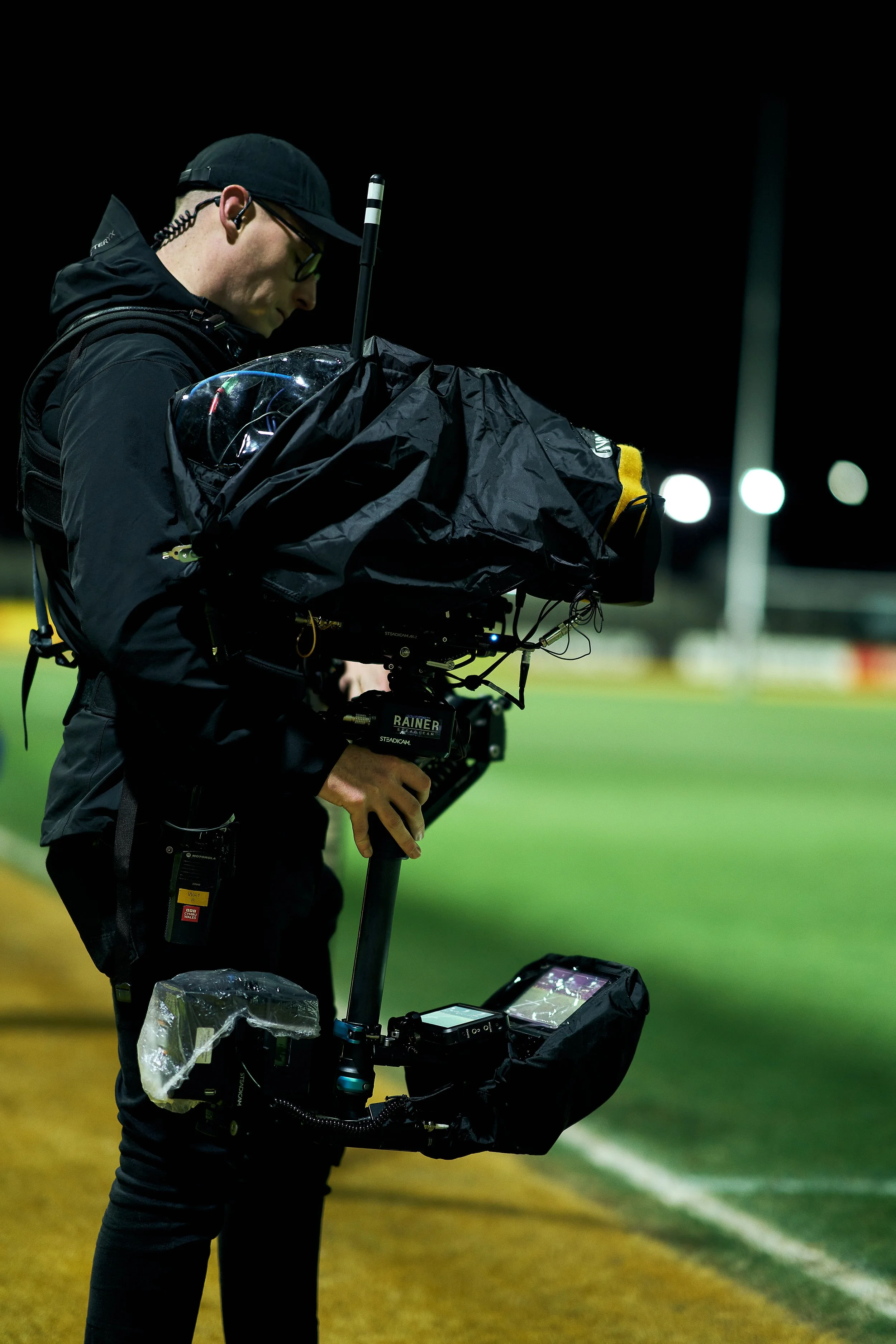 A videographer filming sports game at night on a field, wearing a black jacket, glasses, and a cap, holding a professional camera rig.