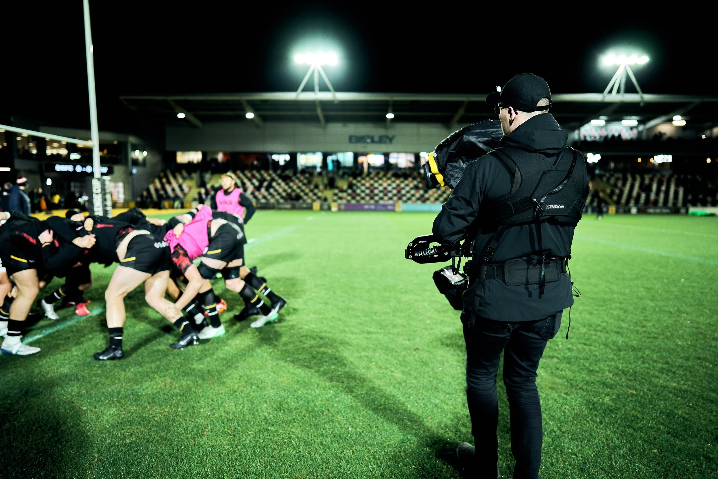 A rugby match taking place at night in a stadium, with players engaging in a scrum and a cameraman filming on the sidelines.