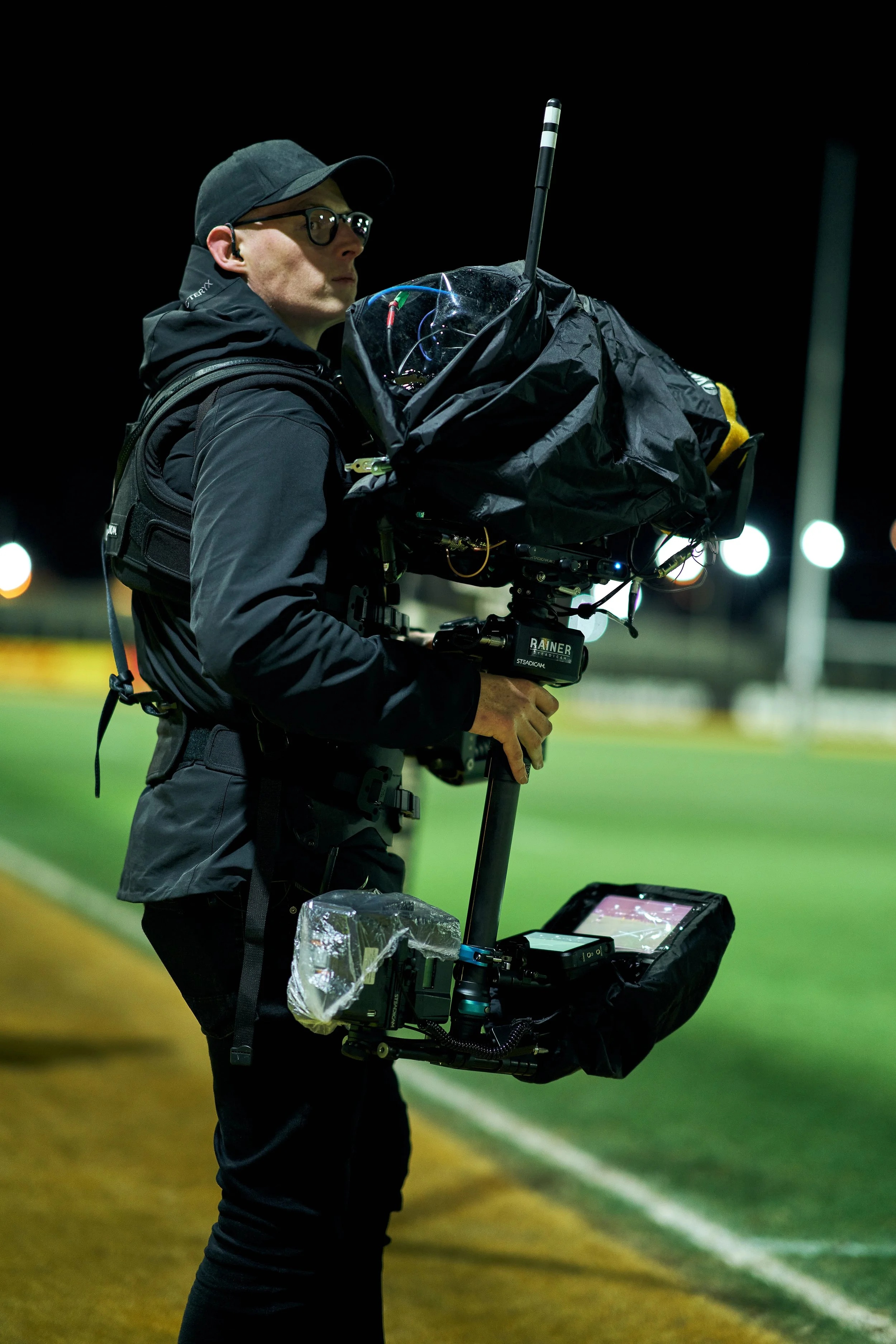 A man standing on a sports field at night, holding a professional camera stabilizer rig, wearing glasses, a black jacket, and a black cap, with bright stadium lights in the background.