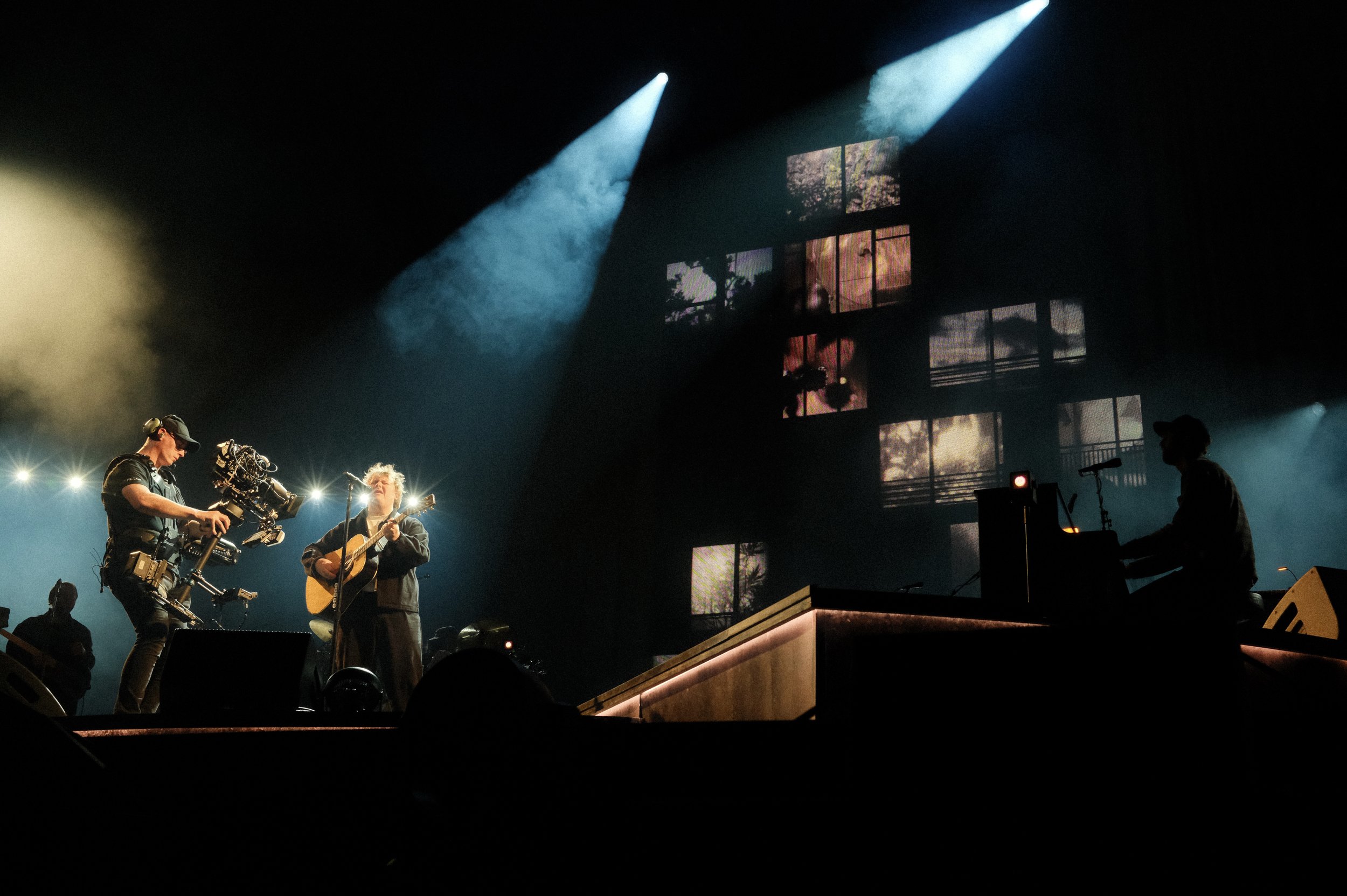Musicians perform on a dimly lit stage with colorful lights, including a guitarist, a singer, and a keyboard player at a concert.