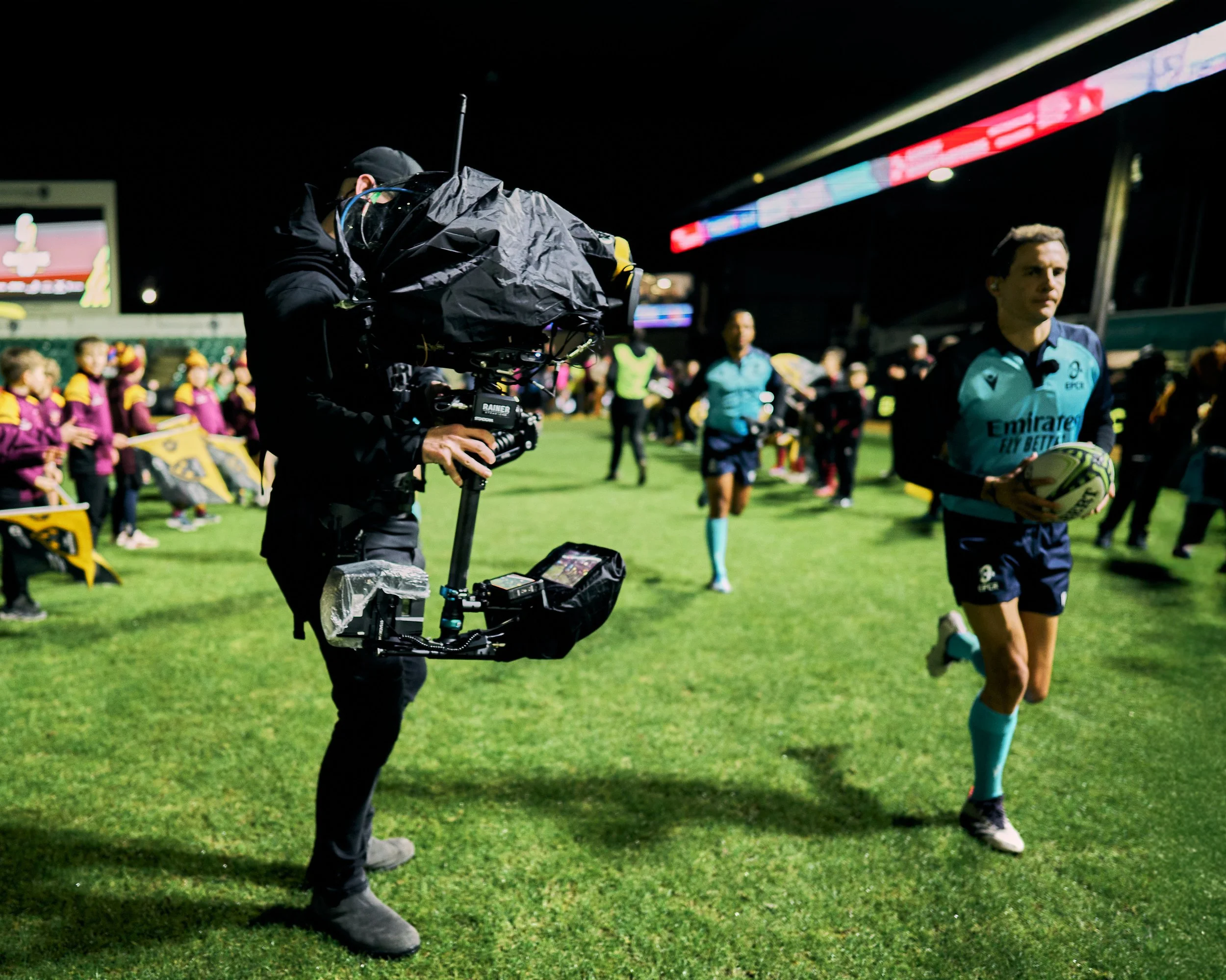 A rugby player in a teal uniform holding a rugby ball while running on a field at night, with a group of children and spectators in the background, and a cameraman recording the scene.