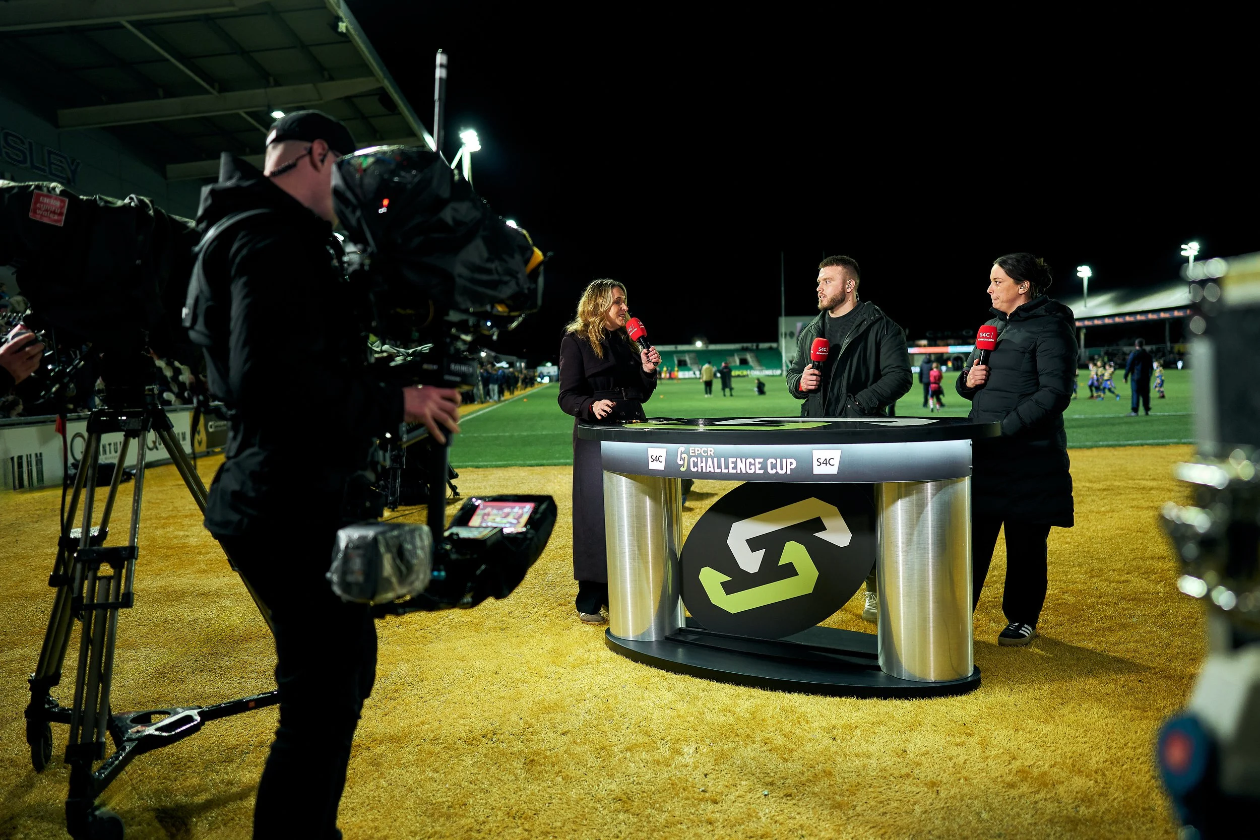 Sports interview taking place on a football field at night with three journalists with microphones, a woman in a black coat, a man in a black jacket, and a woman in a black jacket, behind a table with a tournament logo, while a cameraman records the 