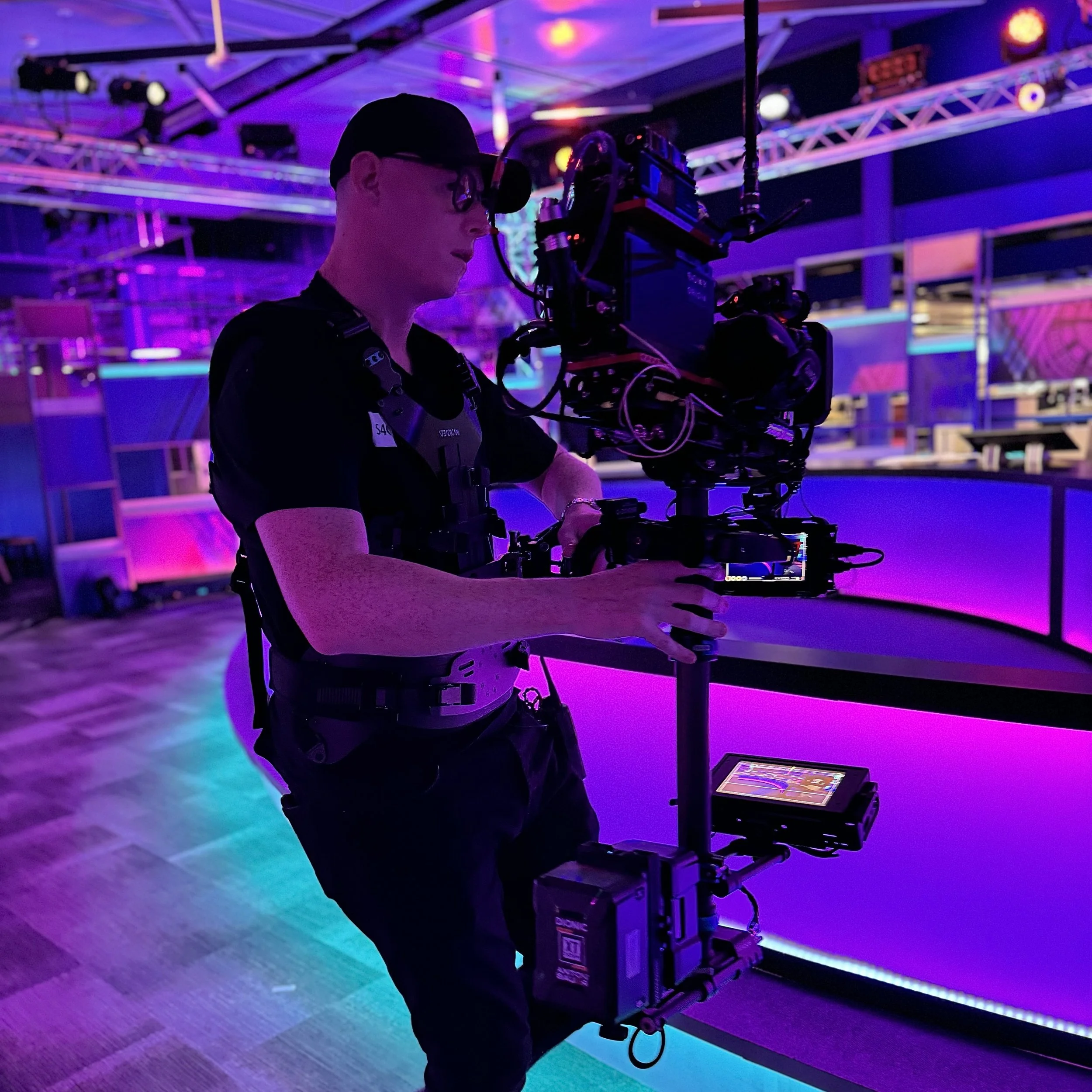 A videographer operating a professional camera on a stabilizer rig in a vibrant, neon-lit venue with purple and pink lighting.