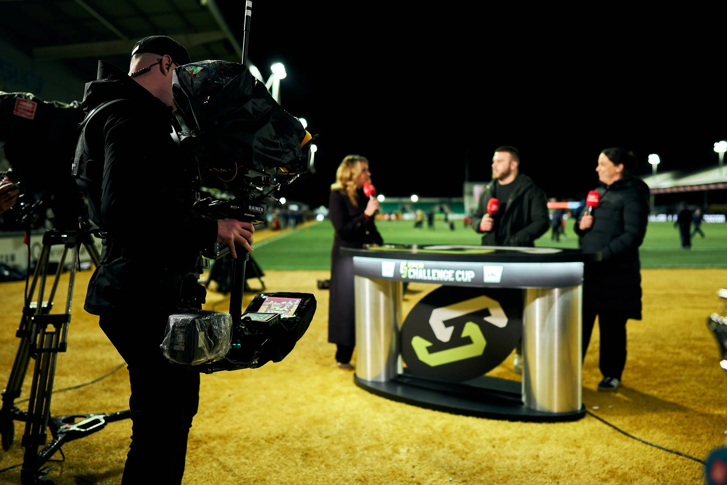 Nighttime sports broadcast setup with a camera operator filming a televised interview at a cricket or baseball field, including three reporters standing at a desk with microphones, and a crowd in the background.
