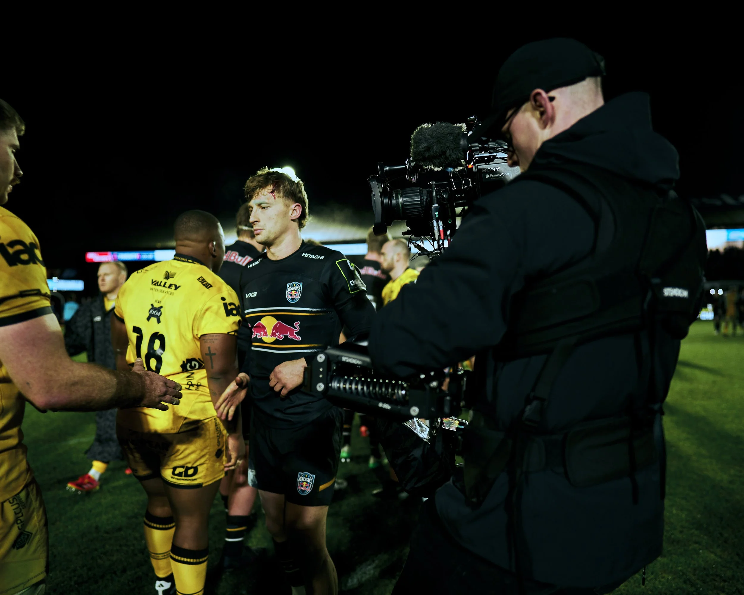 A soccer player in a black jersey with a Red Bull logo talking to players in yellow jerseys during a nighttime match, with a cameraman filming the scene.