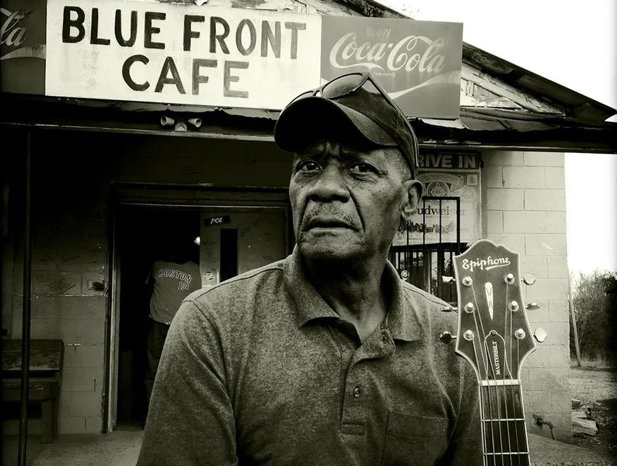 A man in a cap and polo shirt stands in front of Blue Front Cafe with a guitar headstock visible. The cafe has a Coca-Cola sign. The image is in black and white.