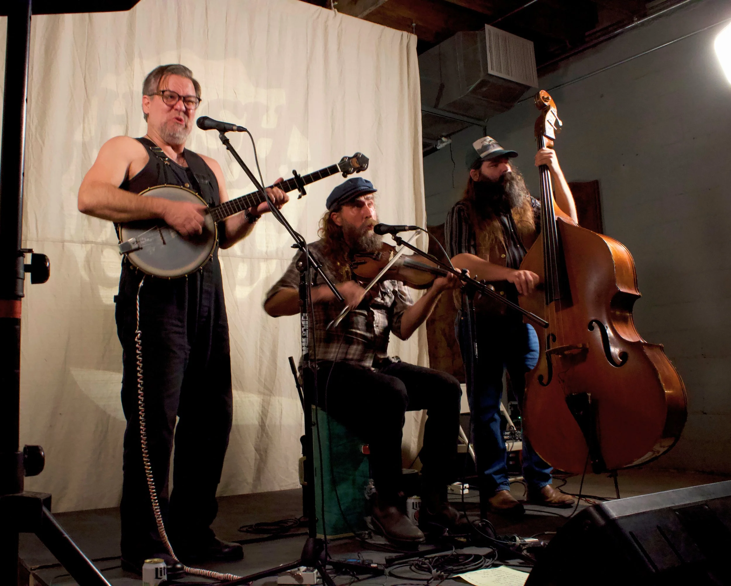 Three musicians performing on a small stage with a plain light-colored backdrop. The man on the left is playing a banjo, the man in the middle is playing a violin, and the man on the right is playing a double bass.