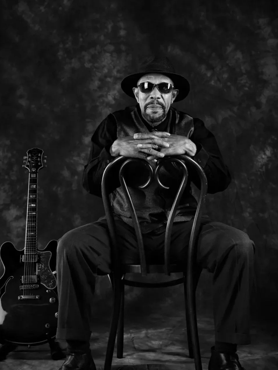 Black and white photo of an older man wearing sunglasses and a hat, sitting on a chair with his hands resting on the backrest, beside an electric guitar. The man is dressed in dark clothing and the background is blurred.