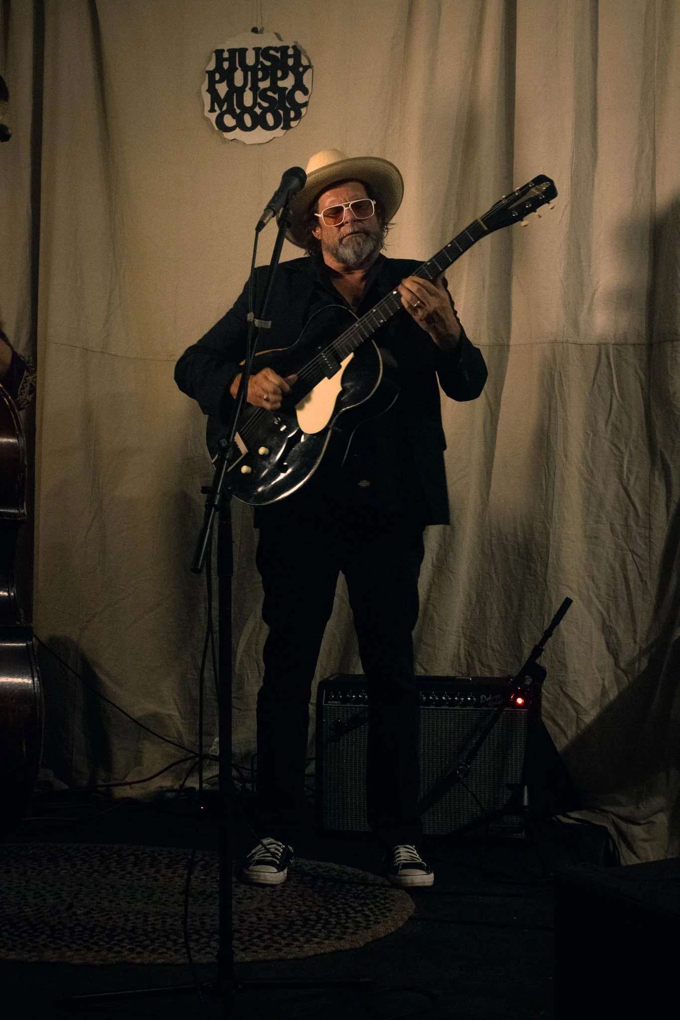 A man with a beard, wearing sunglasses, a wide-brimmed hat, and a black shirt, is playing an electric guitar on stage. There is a microphone in front of him and a small amplifier on the floor. Behind him is a curtain, and a sign that reads 'Hush Puppy Music Coop' is on the wall.