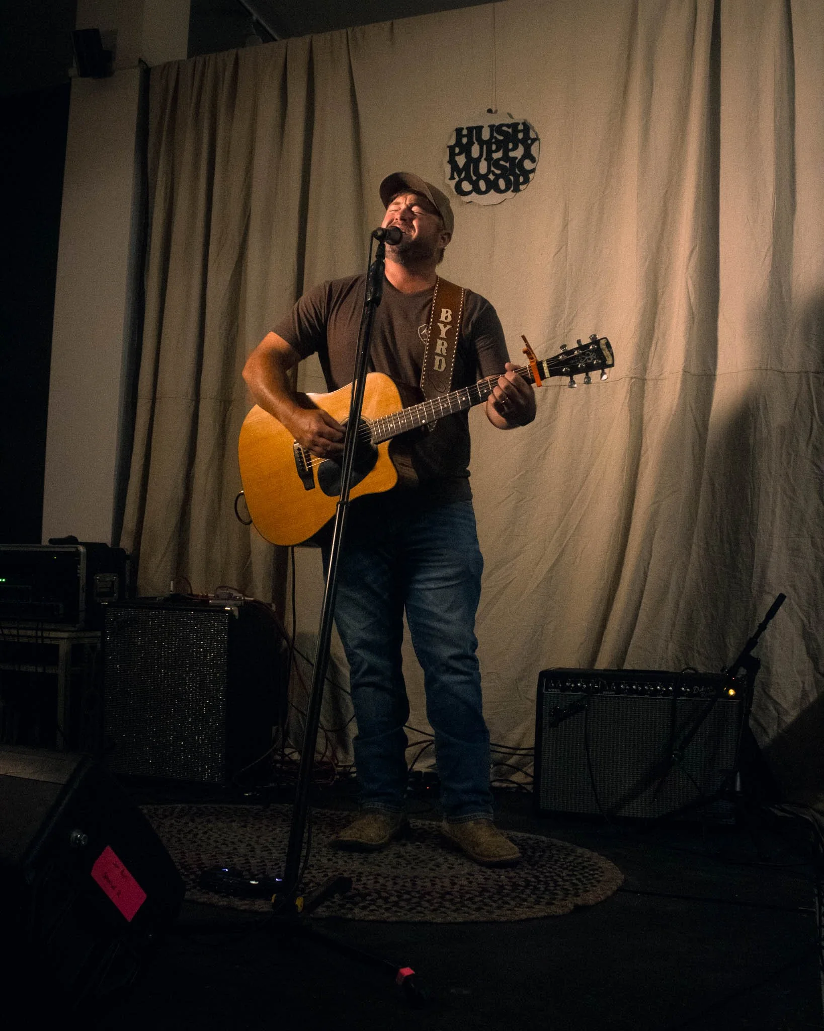 Male musician singing and playing an acoustic guitar on stage, with sound equipment and a curtain in the background.