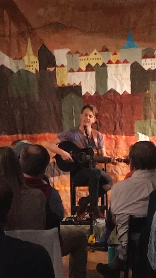 Musician playing acoustic guitar on stage with colorful backdrop in front of seated audience.