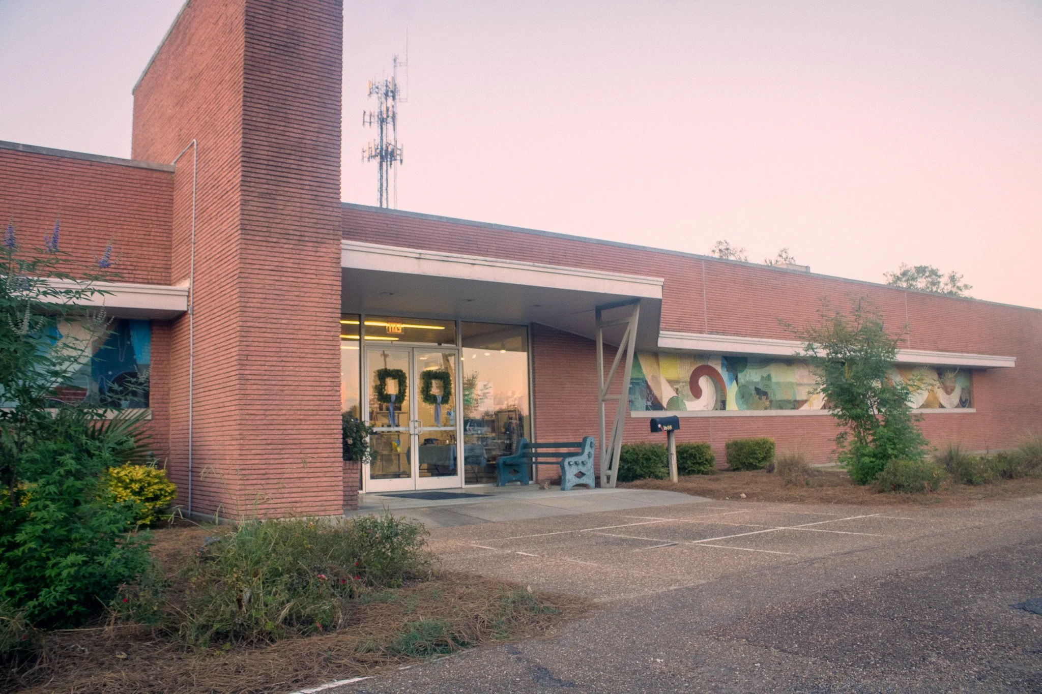 Exterior of the Museum of the Mississippi Delta in Greenwood, MS, featuring a red brick facade, glass entrance doors, a colorful mural along the right wall, and a parking area in the foreground under a twilight sky.