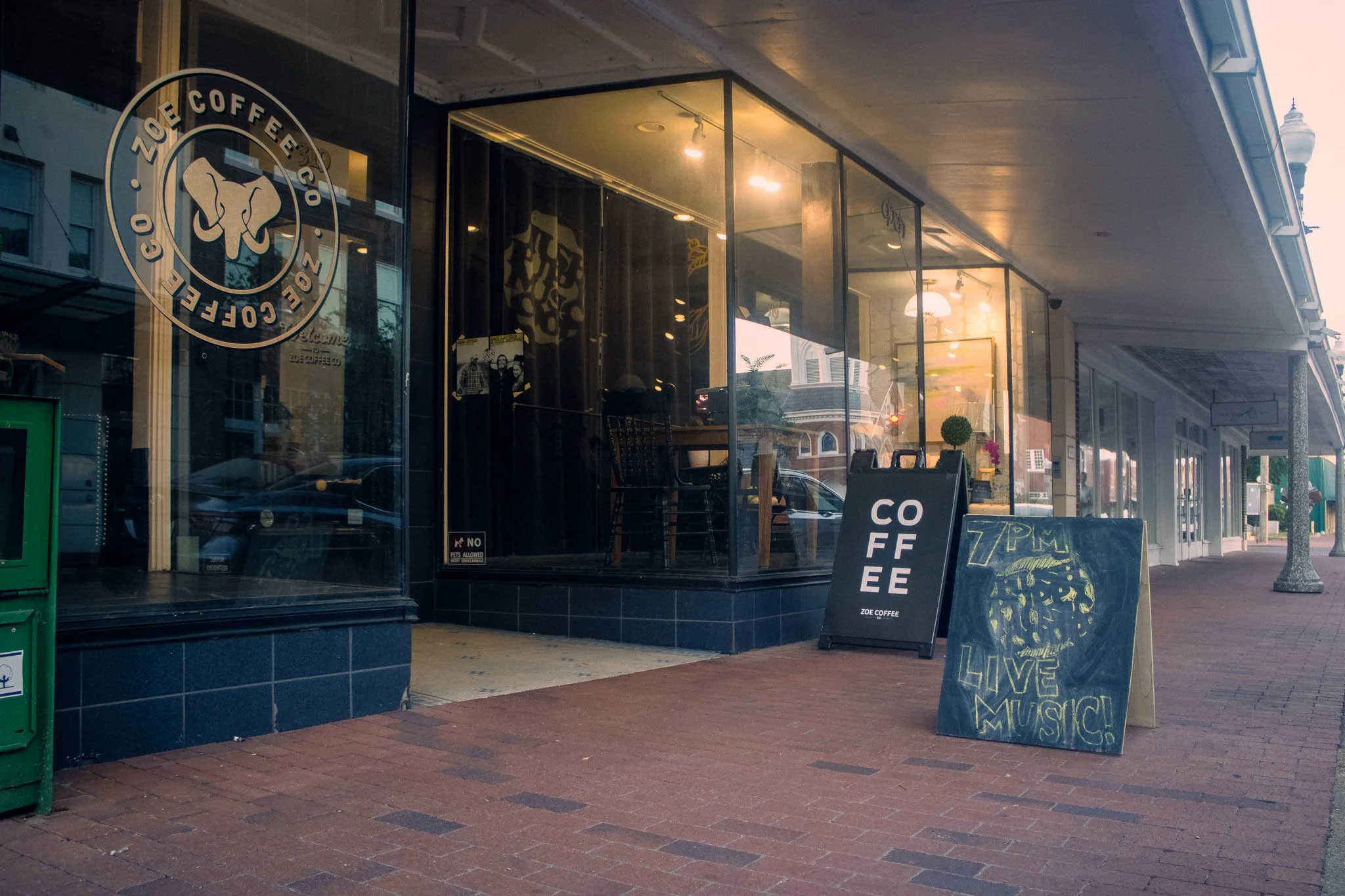 The exterior storefront of Zoe Coffee Co. in Greenwood, MS, featuring large glass windows, a circular logo, and a brick sidewalk. A chalkboard sign on the sidewalk reads "7PM Live Music!".