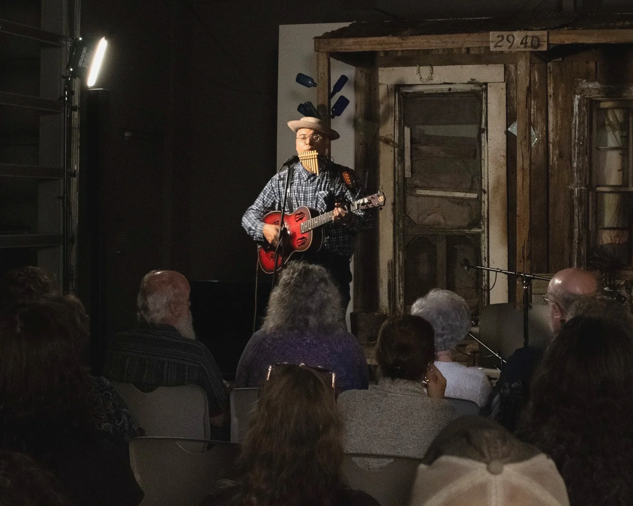 A musician wearing a hat and glasses is playing a guitar and singing into a microphone during a performance. The setting appears to be a rustic indoor venue with dim lighting.
