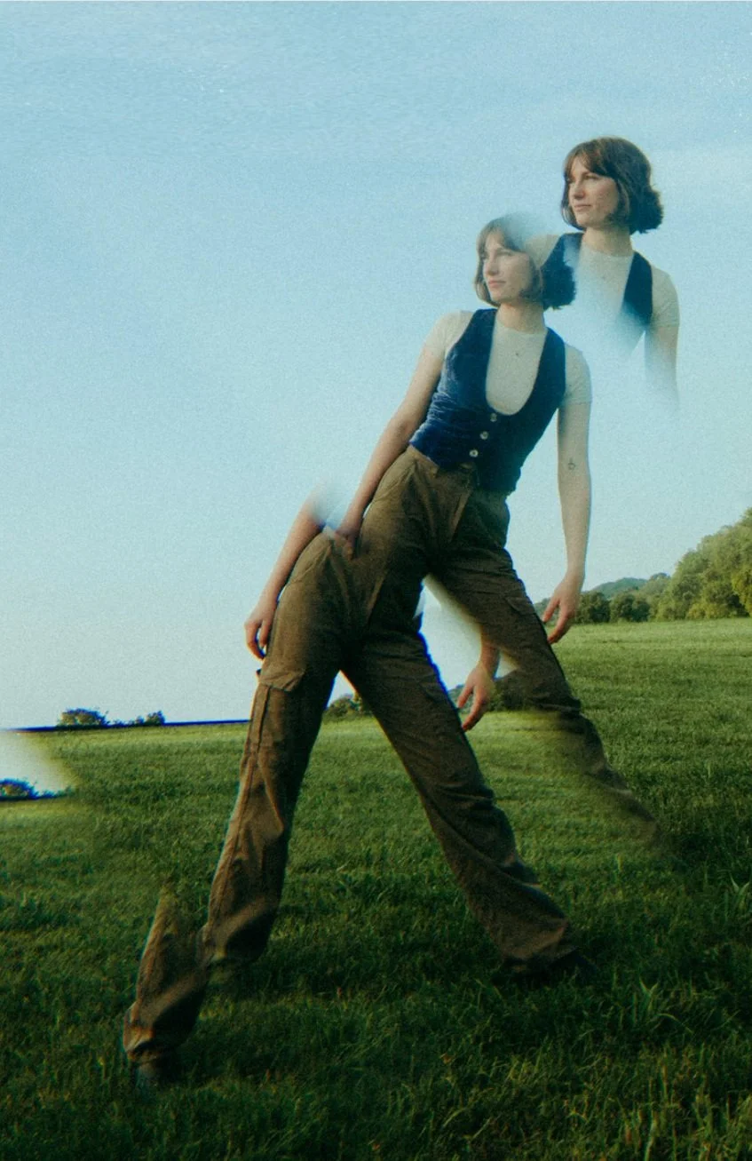 A double-exposure portrait of indie-folk musician Remi Goode standing in a sunlit grassy field, creating a dreamy, cinematic effect.