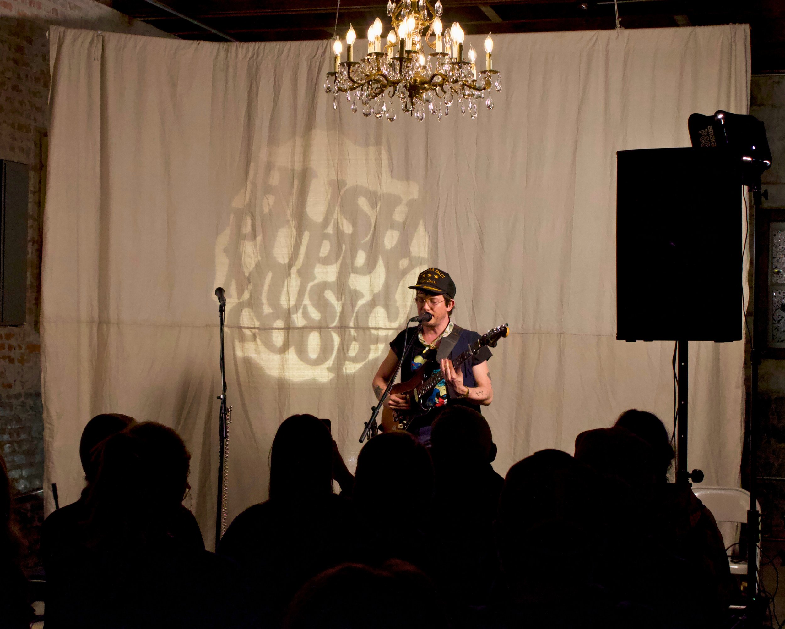 A musician performs on stage with a guitar and microphone, audience members seated in front, a chandelier hanging above, and a backdrop with a projected shadow pattern.