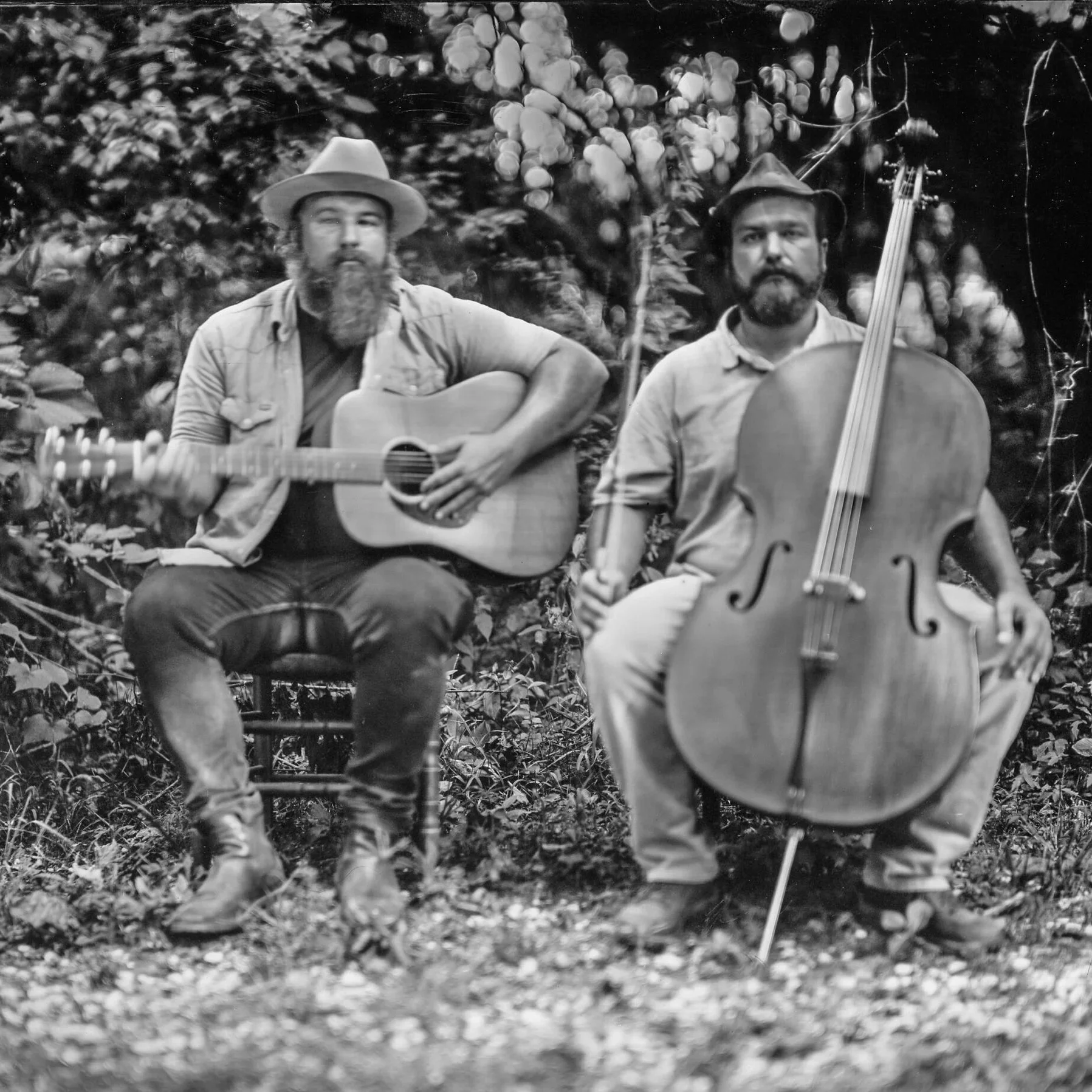 Two men sitting outdoors, one playing an acoustic guitar and the other playing a double bass.