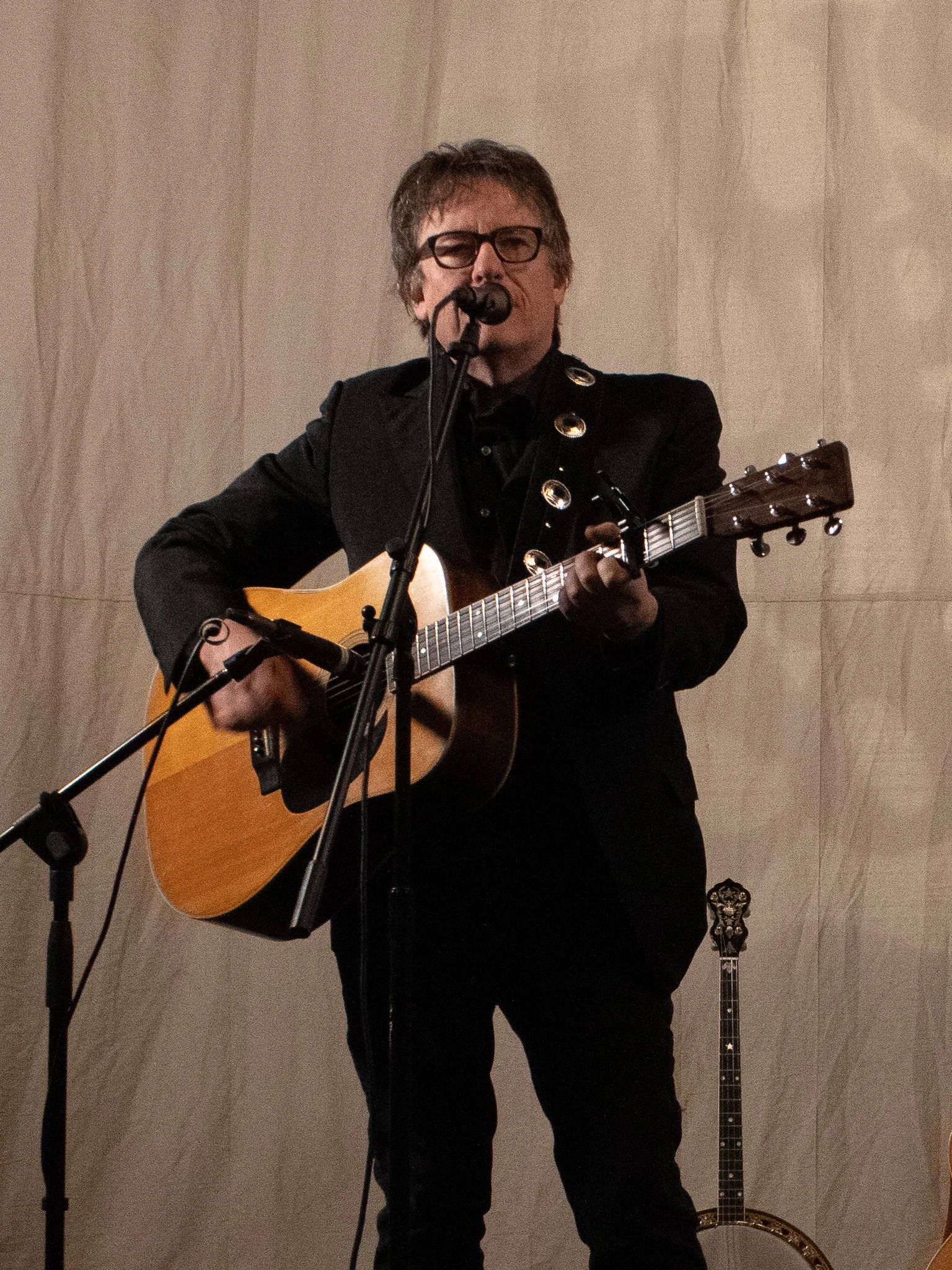 A man with glasses and brown hair is playing an acoustic guitar and singing into a microphone on stage. He is wearing a black suit and is standing in front of a light-colored background. There is another guitar visible in the background.