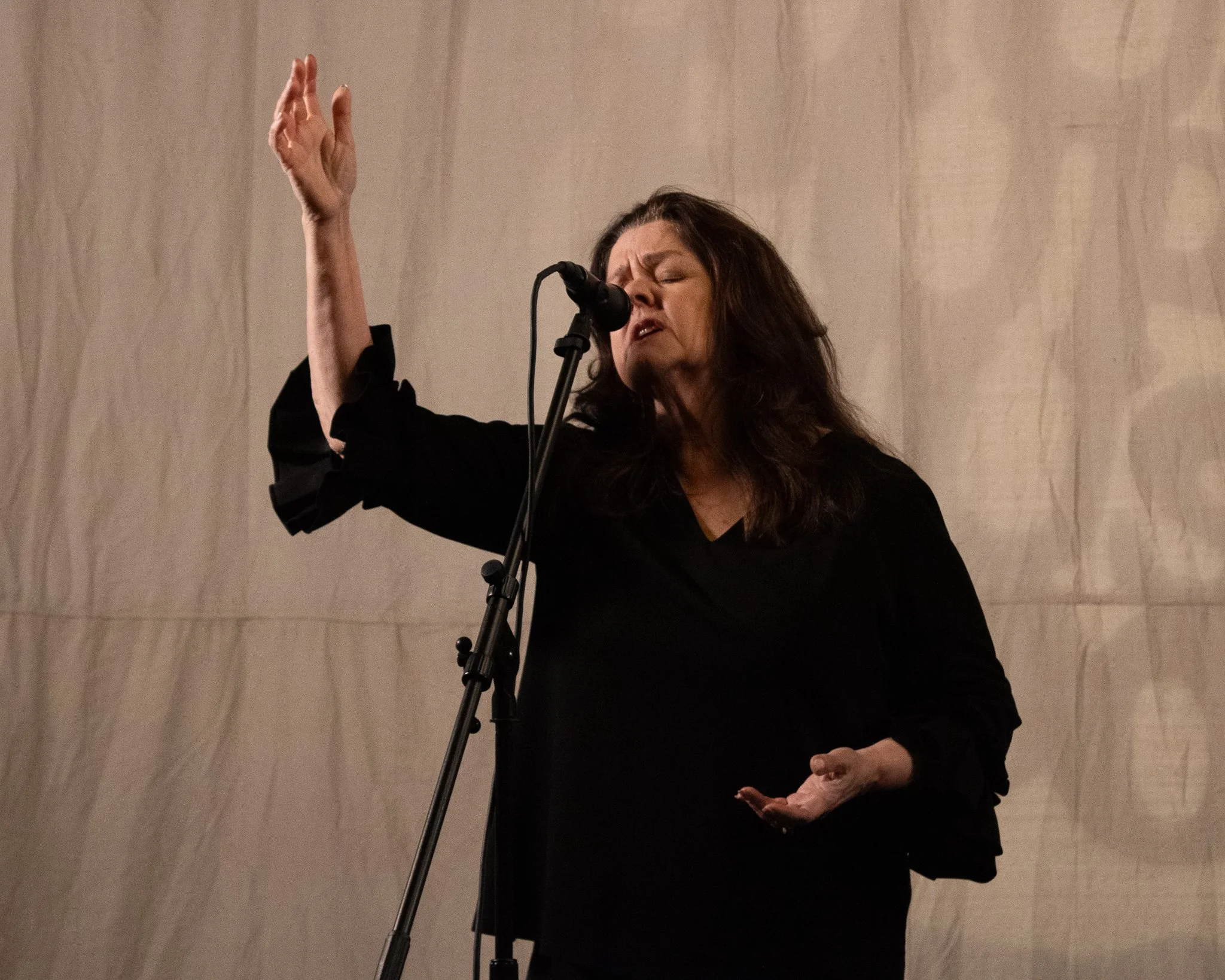 A woman singing passionately into a microphone, with one hand raised and the other hand near her waist, against a plain, textured background.
