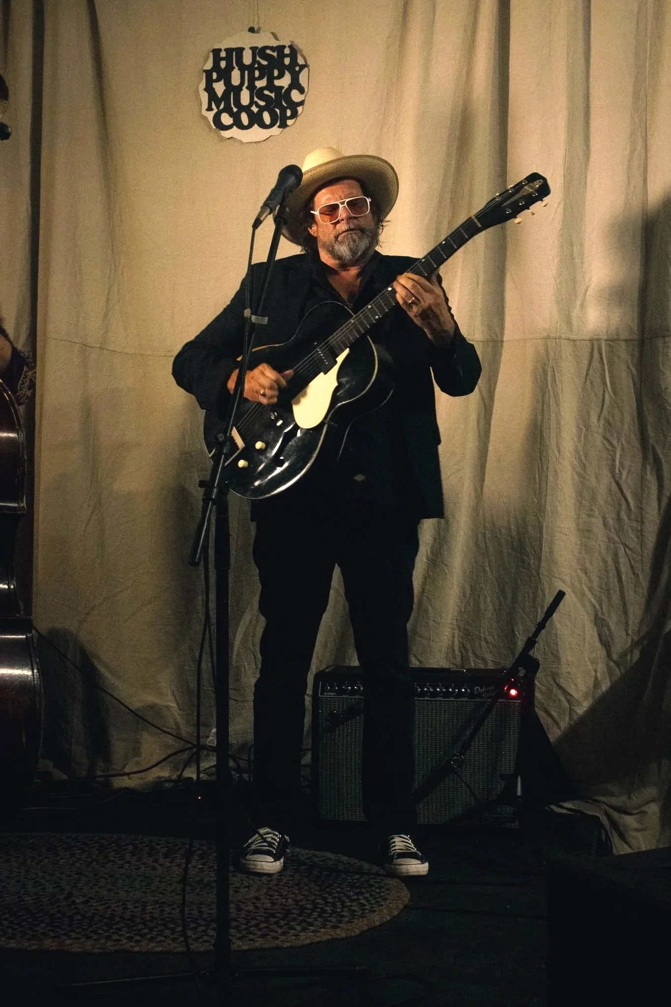 A man with sunglasses and a hat playing an acoustic guitar on stage, with a sign above that reads 'Hush Puppy Music Coop' behind him.