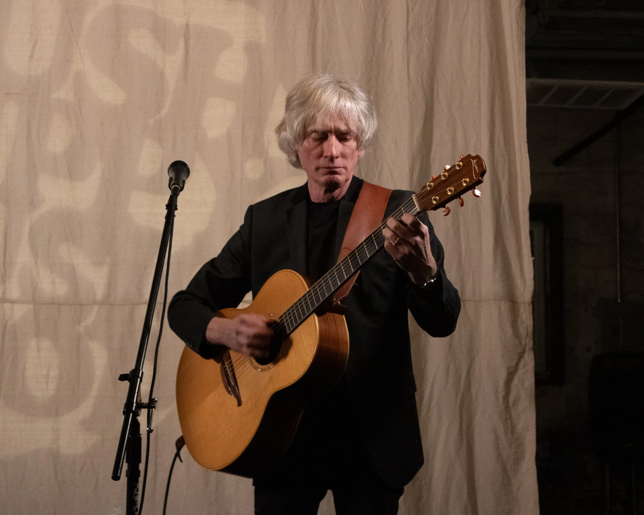 Man with gray hair playing acoustic guitar on stage with beige backdrop.