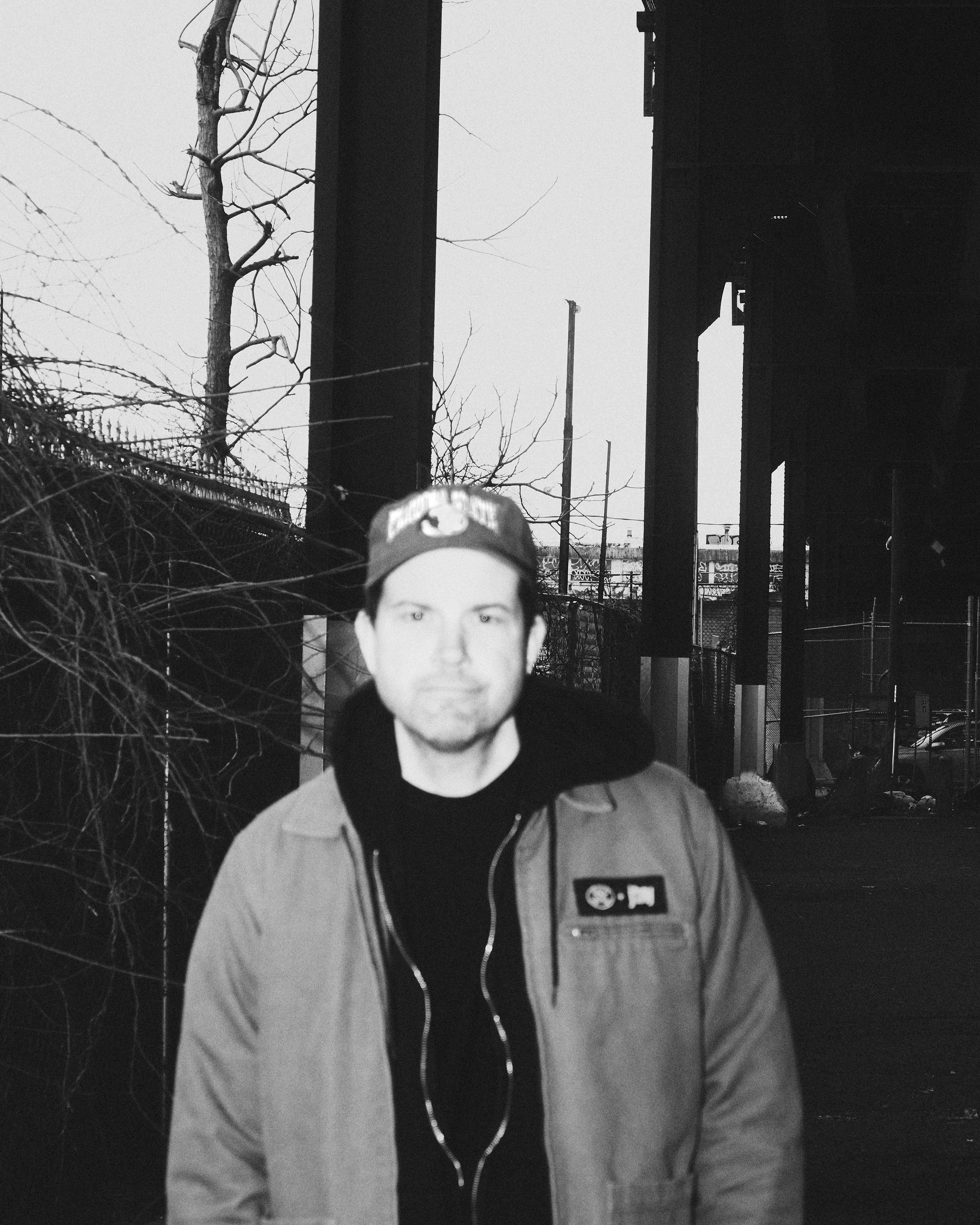 Black and white portrait of John Ross, frontman of the indie rock band Wild Pink, standing outdoors under an overpass wearing a cap and jacket.