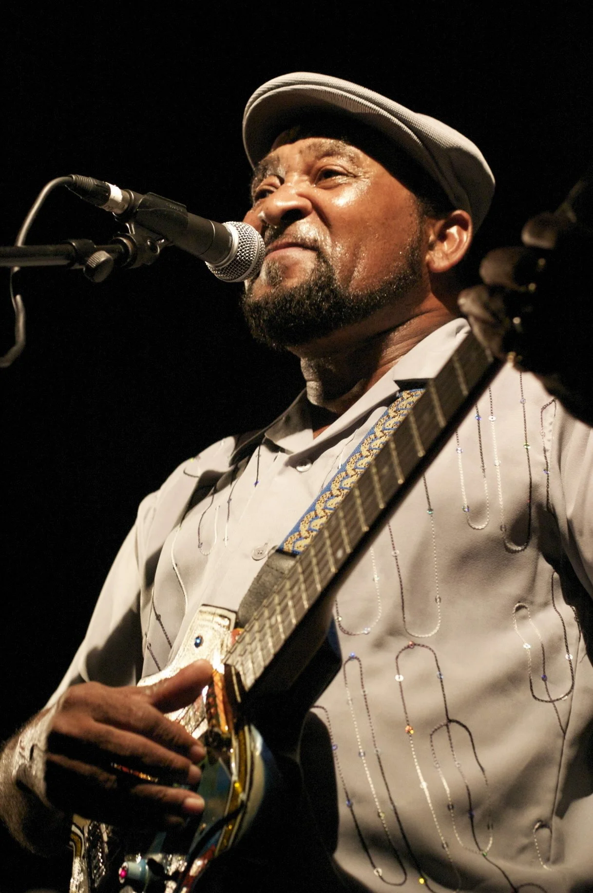 A man singing into a microphone while playing an electric guitar, wearing a light-colored shirt with decorative patterns and a gray hat.