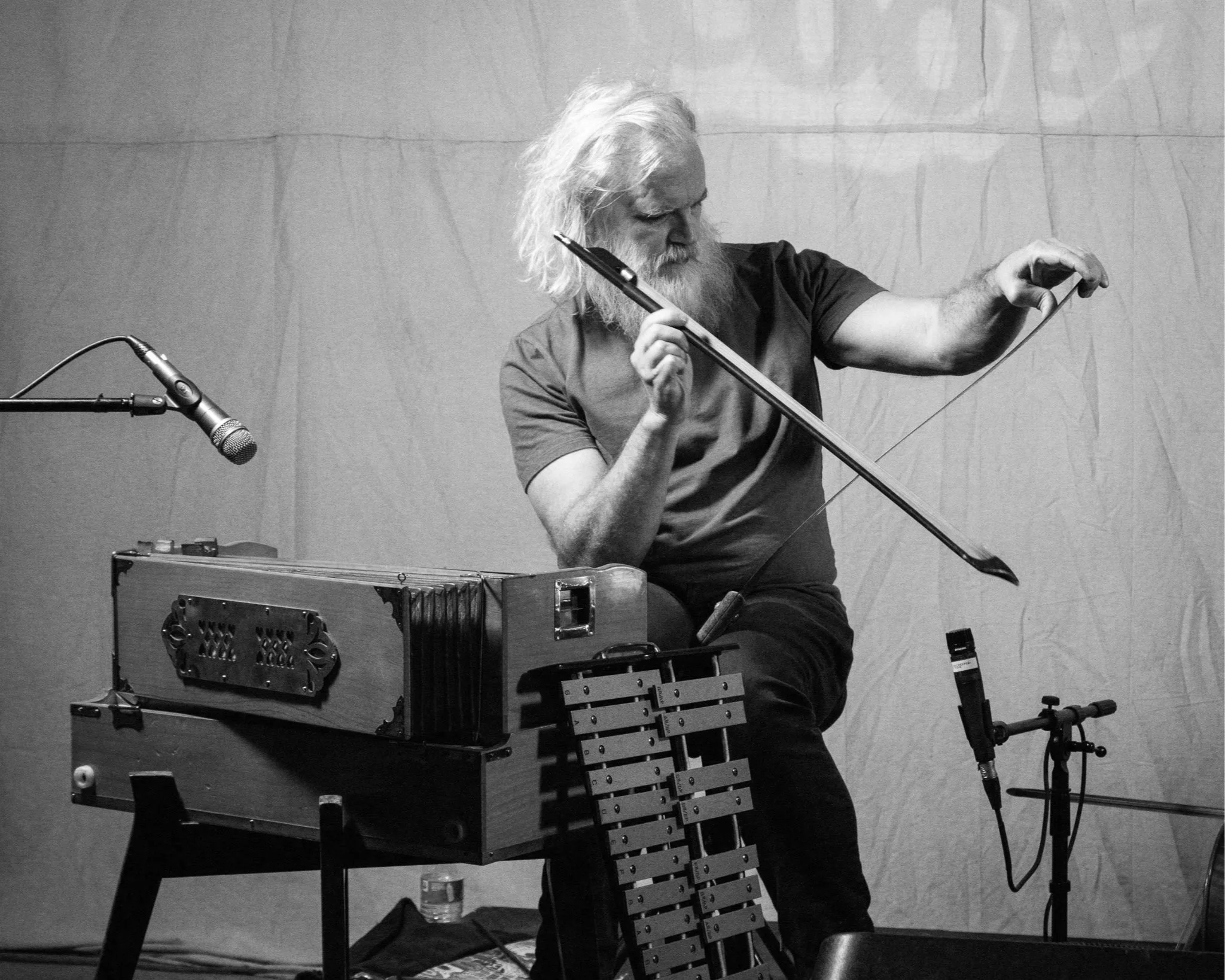 An elderly man with long white hair and a beard playing a cello. There are microphones and electronic equipment around him, and he is standing in front of a plain wall.