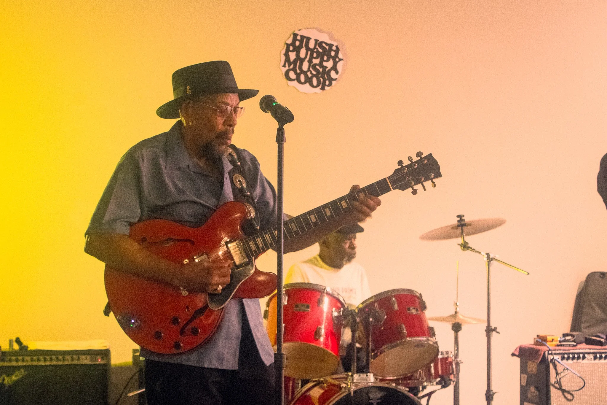 A man wearing a black hat and glasses playing an electric guitar on stage, with a drum set behind him, during a live music performance.