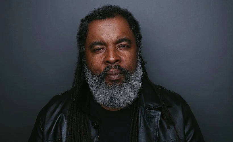 A man with long hair and a gray beard wearing a black leather jacket against a dark background.