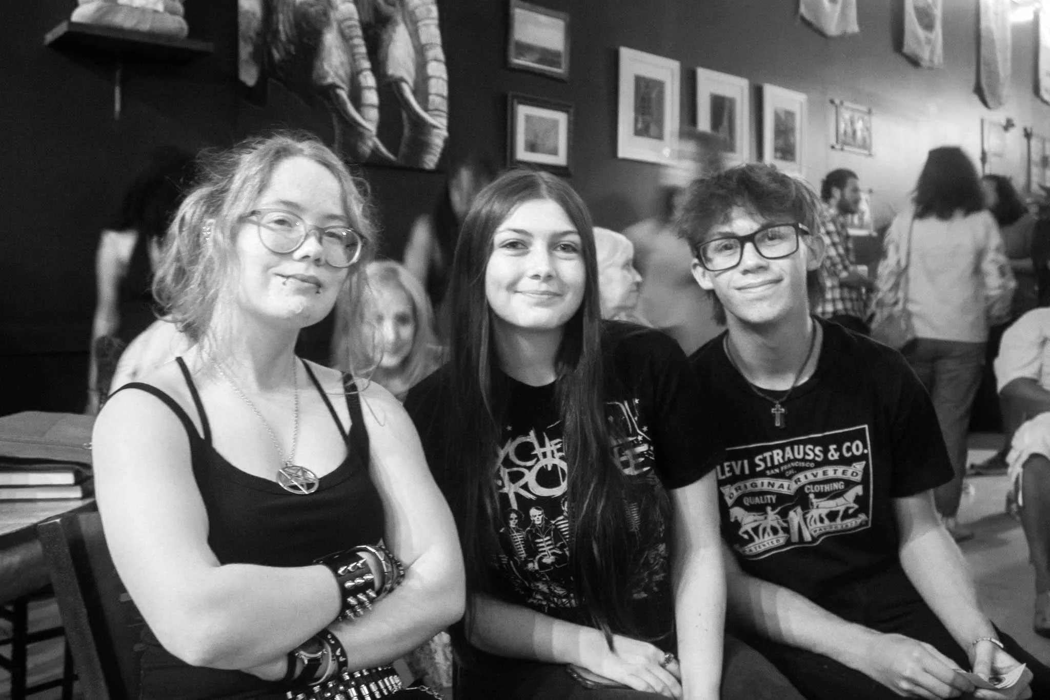 Three young people sitting at a table in a social setting, smiling and facing the camera, with art and photographs on the wall behind them, and other people in the background.