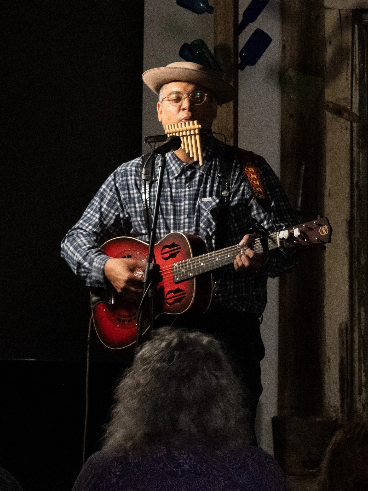 A musician wearing glasses, a hat, and a plaid shirt plays an acoustic guitar, with a pan flute in his mouth, standing in front of a dark background and an audience.