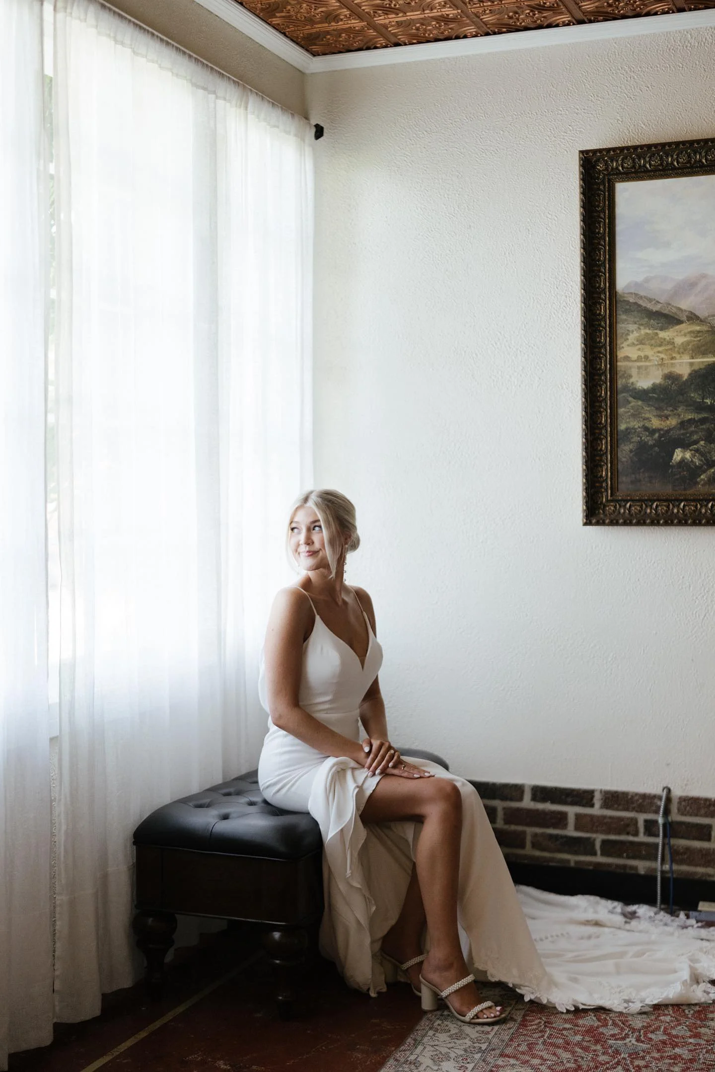 A woman in a white dress sitting on a black bench by a window with white curtains, in a room with a painting on the wall.