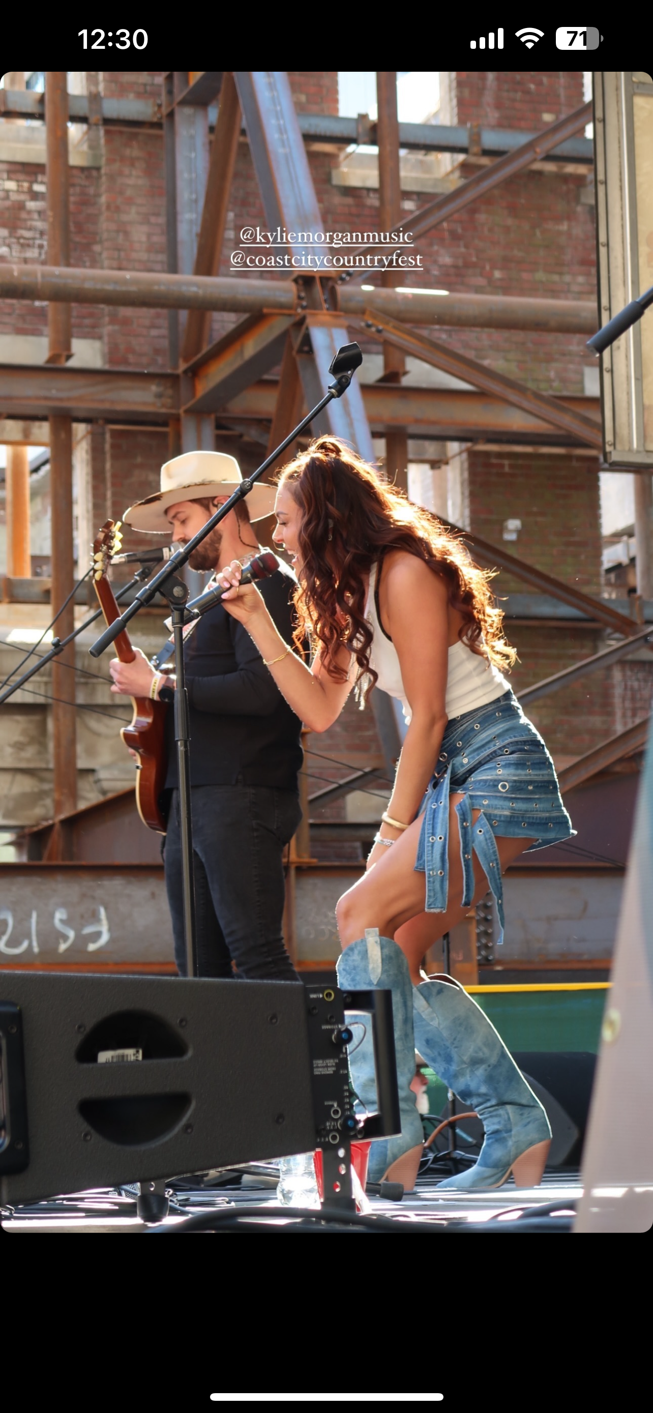 A woman singing into a microphone and a man playing an acoustic guitar on a stage outdoors with a wooden and brick background.