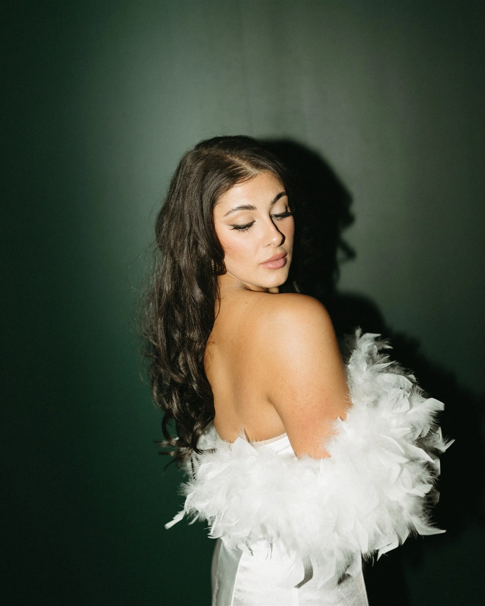 A woman with long dark hair and makeup, wearing a white feathered garment, standing against a dark background.