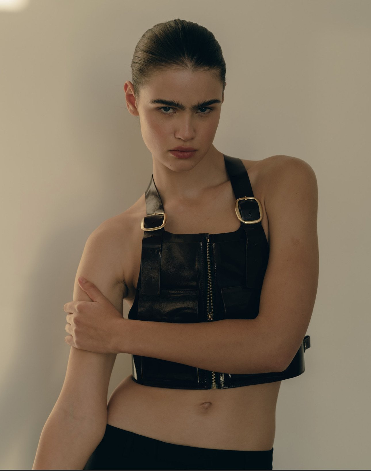Young woman with dark hair in a slicked-back style wearing a black leather halter top with gold buckles, standing against a plain light-colored background with a serious expression.