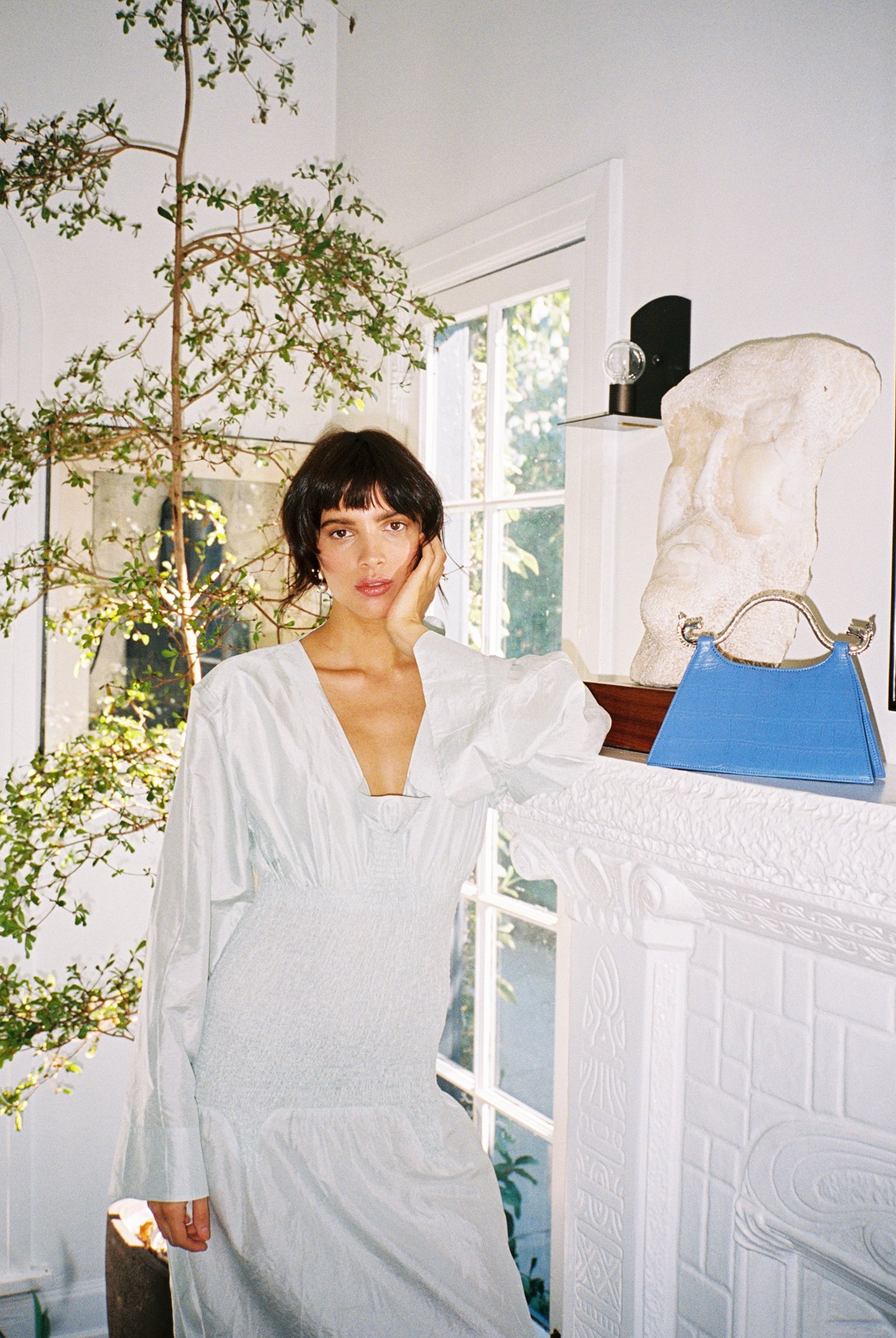 Woman with short dark hair wearing a white dress standing next to a white fireplace with a blue handbag on top. Behind her is a large plant and a window letting in natural light.