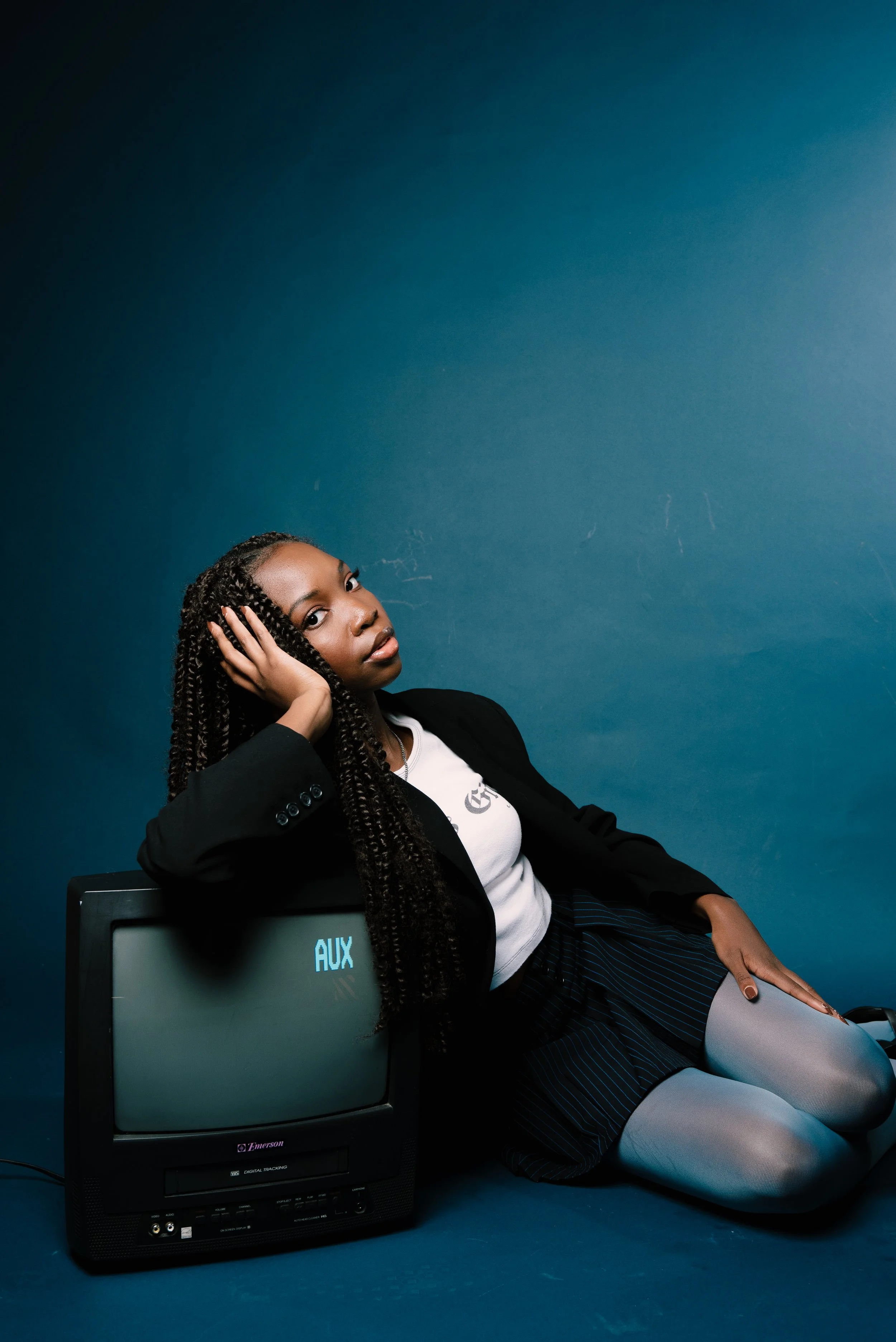 A woman with long twisted hair sitting on the floor, leaning against an old television, with a blue background. She is wearing a black blazer, a white shirt, a black skirt with pinstripes, and light gray tights.